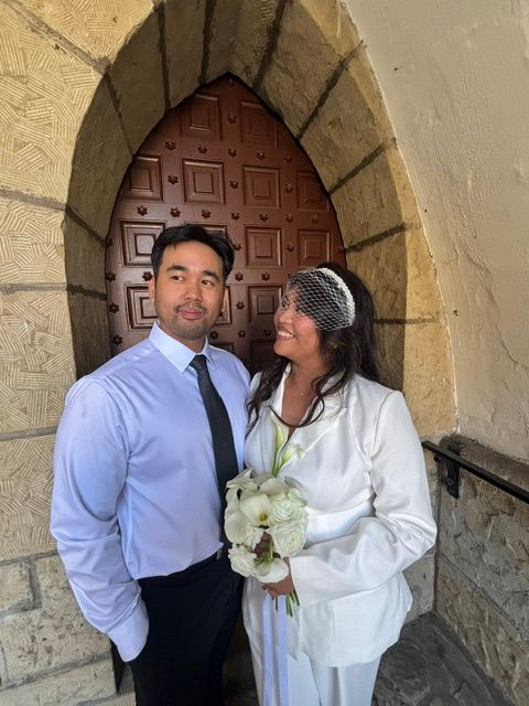 A bride in white holding a bouquet stands next to a groom in a light blue shirt outside a stone building with a wooden door.