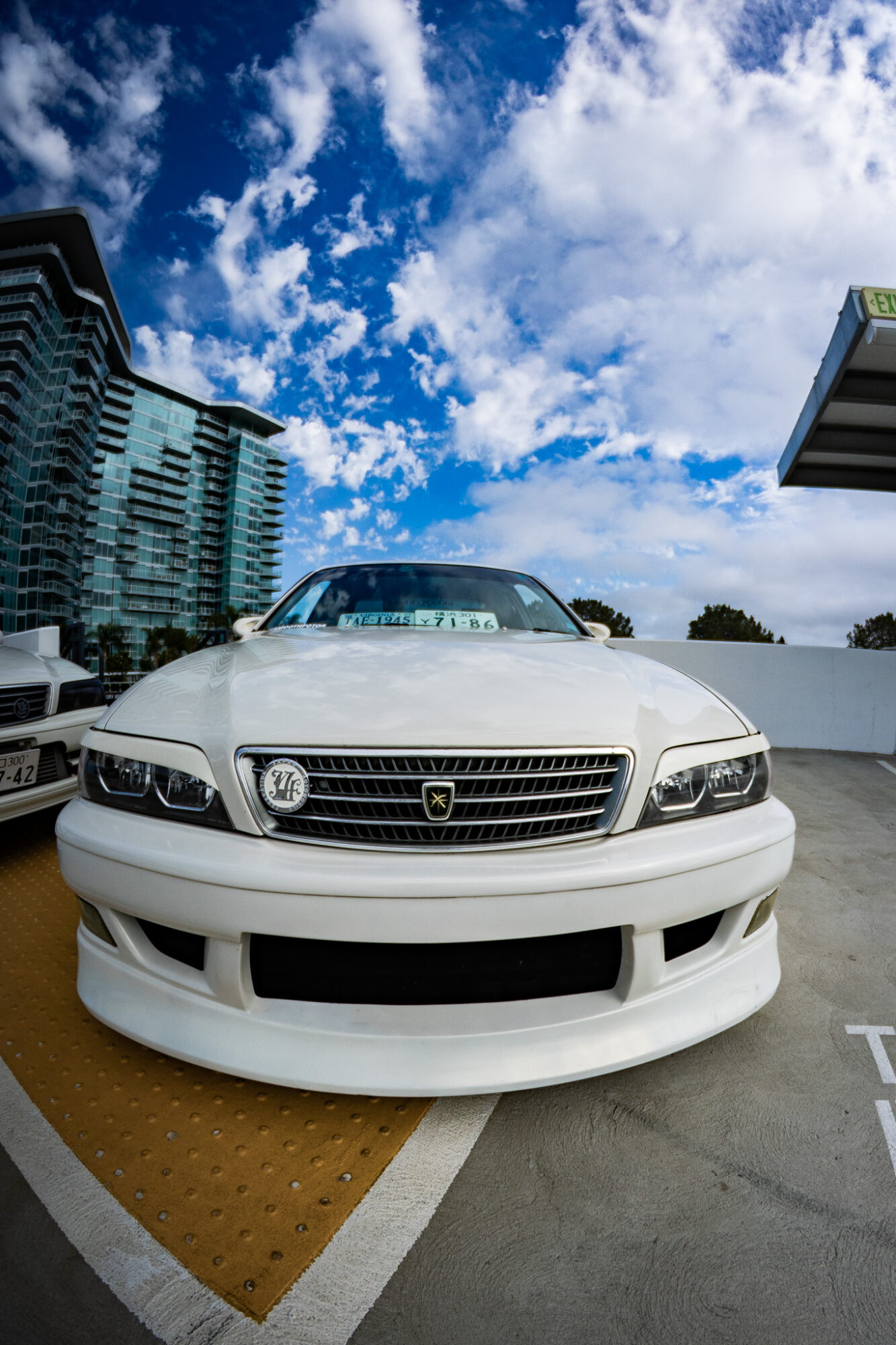 White sports car parked outdoors with modern buildings and a partly cloudy sky in the background.