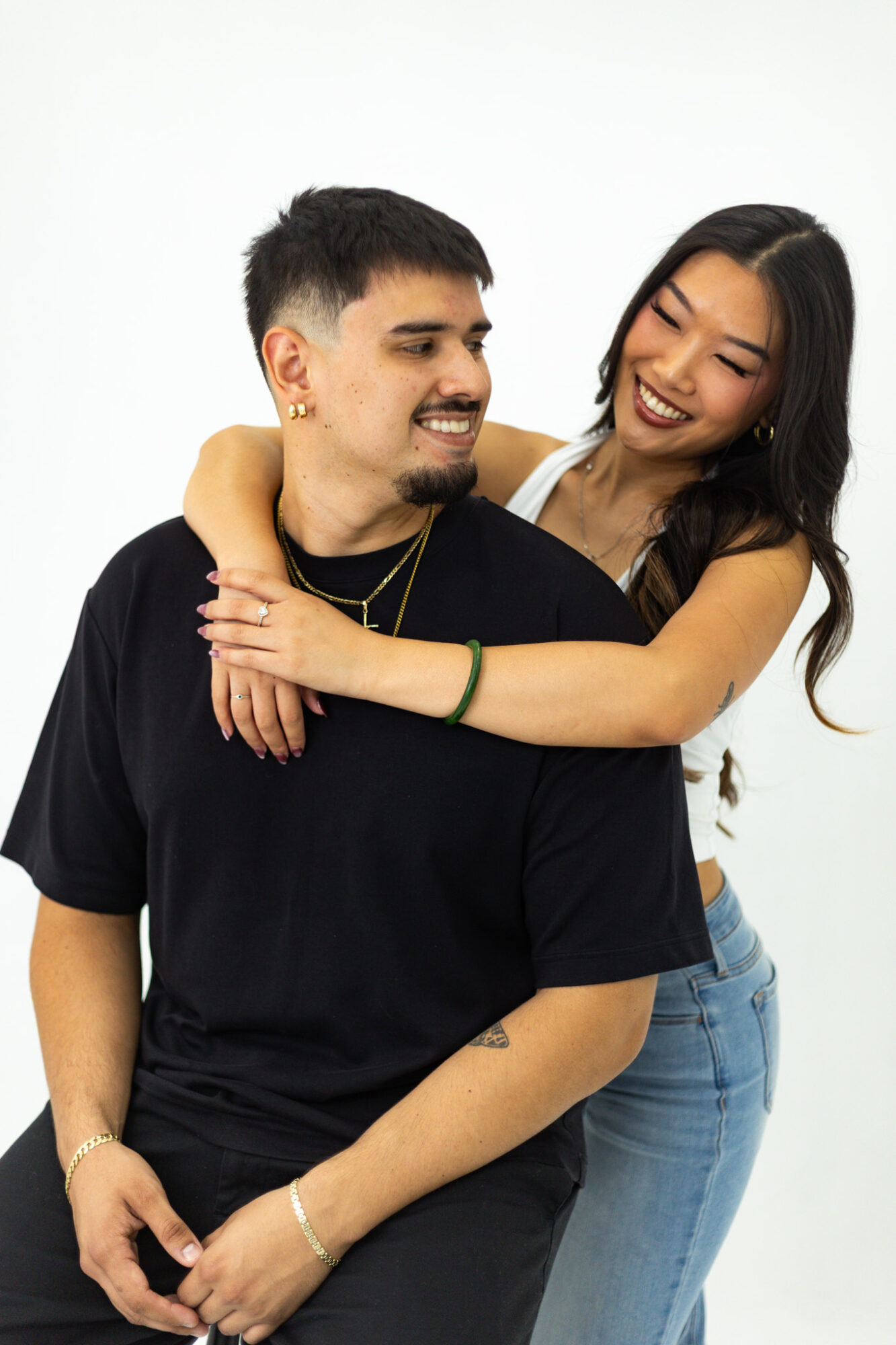 A smiling man and woman embrace, woman wrapping arms around man's shoulders, both looking at each other, against a white background.