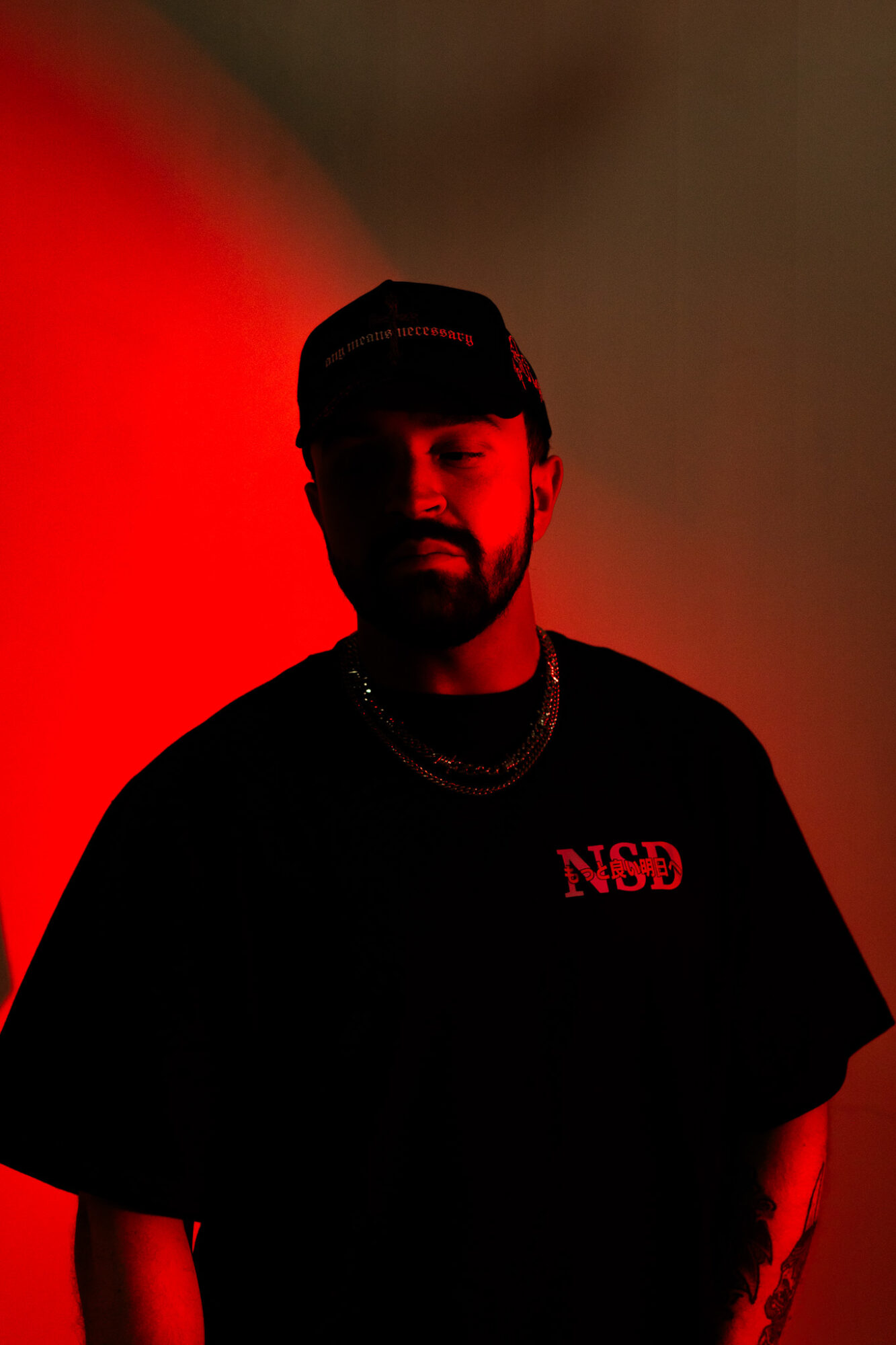 Person with beard and cap, wearing a black shirt with red text, illuminated by red and dark lighting.