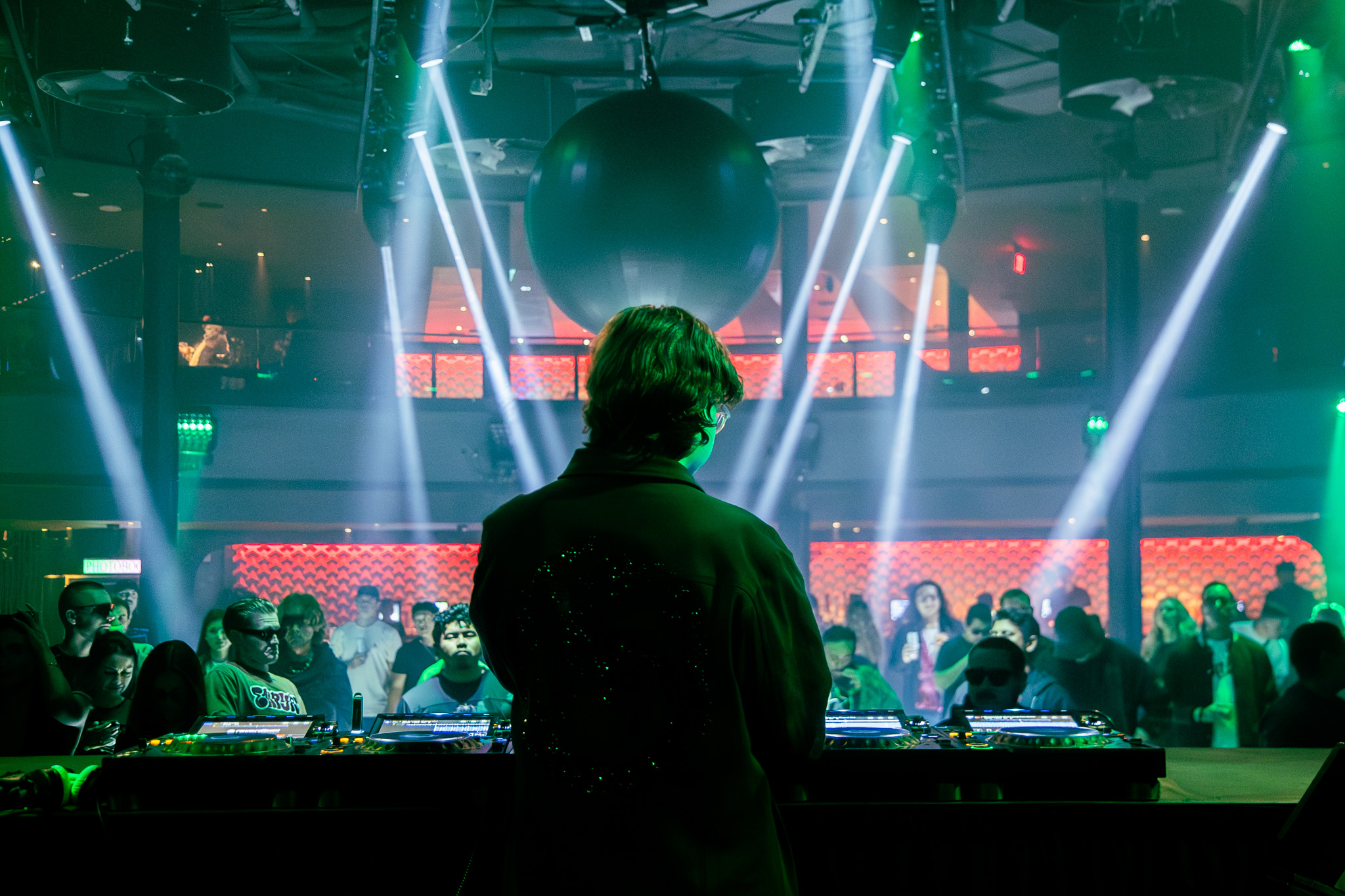 Person with curly hair stands in front of a DJ booth in a nightclub with colorful lights and a crowd.