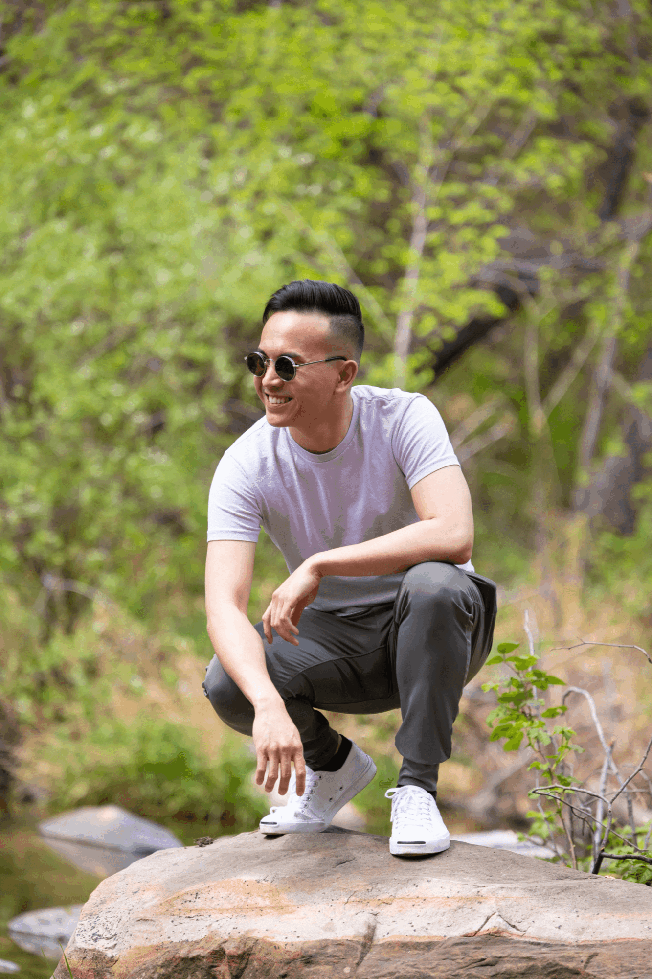 Young man crouching on a rock outdoors, wearing sunglasses, a white t-shirt, and dark pants, with green trees in the background.