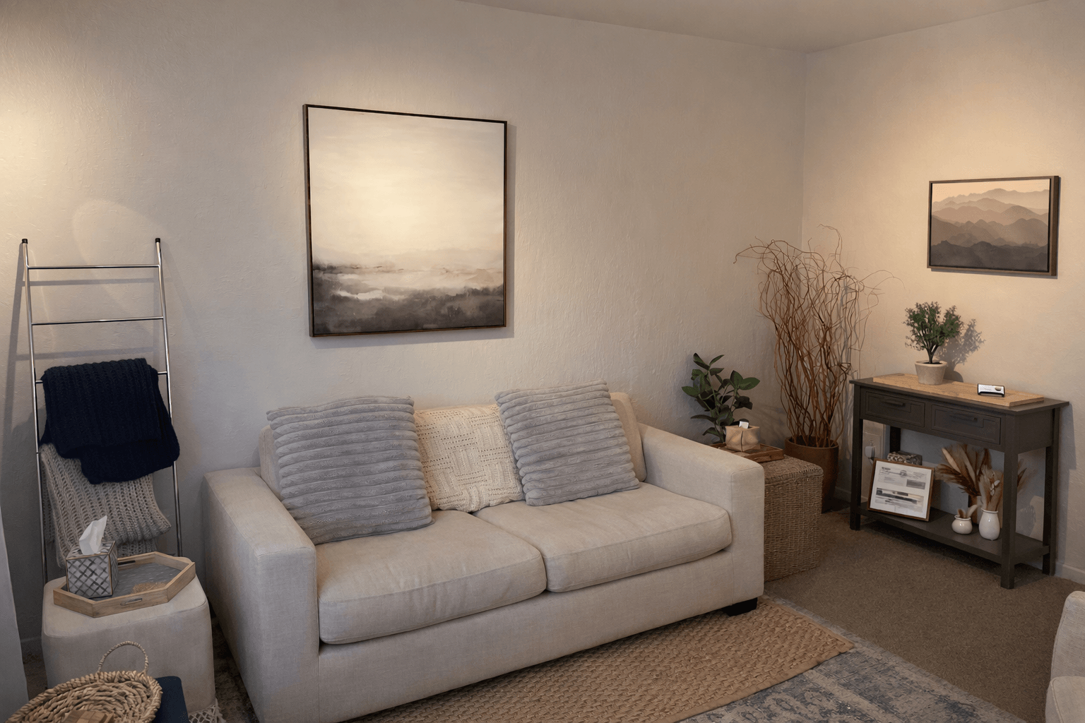 Living room with a white sofa, two gray pillows, wall art, a side table, and a console table with plants and framed items.