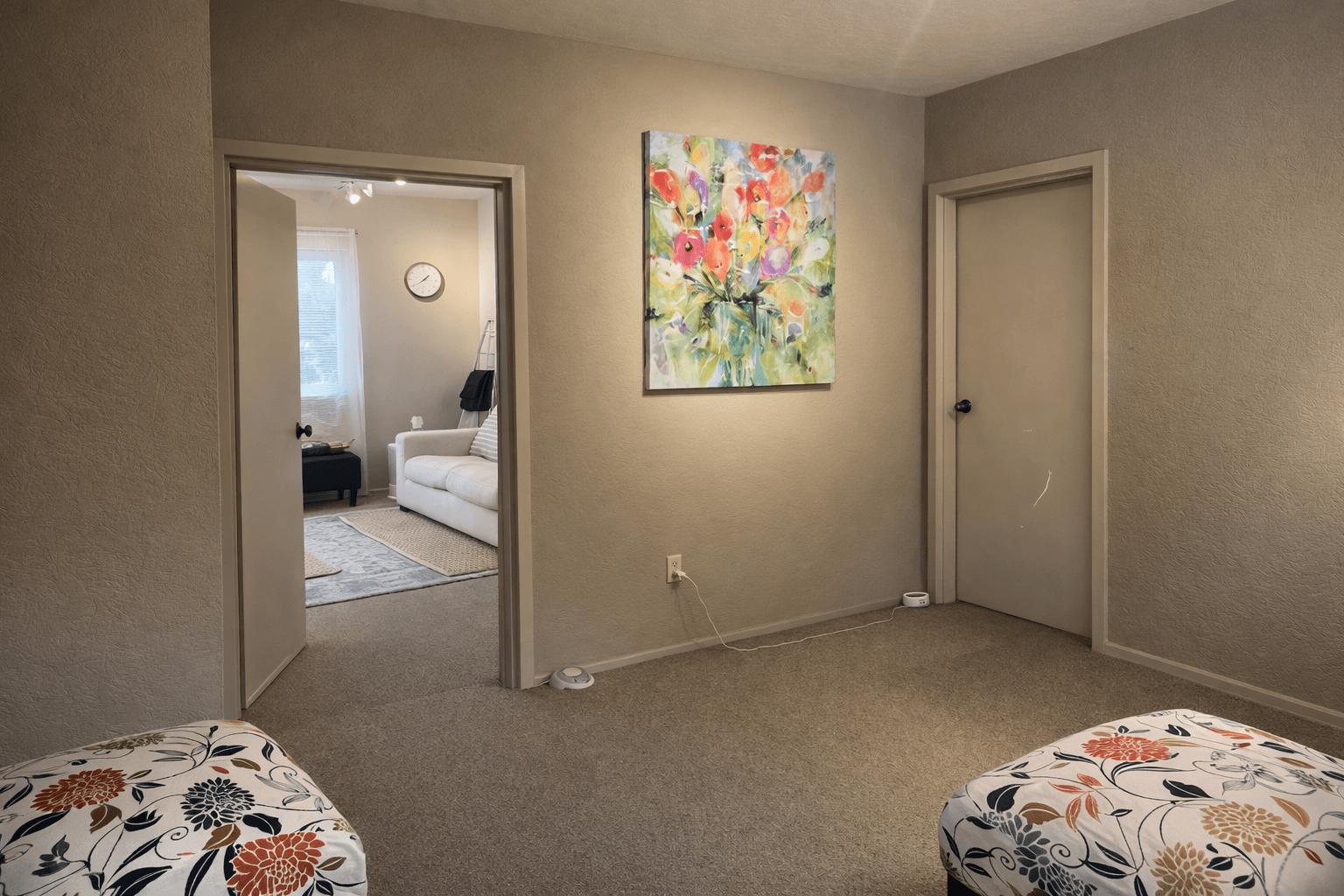Living room with doorway, colorful painting, closed door, and carpeted floor, viewed from a corner with two beds partially visible.