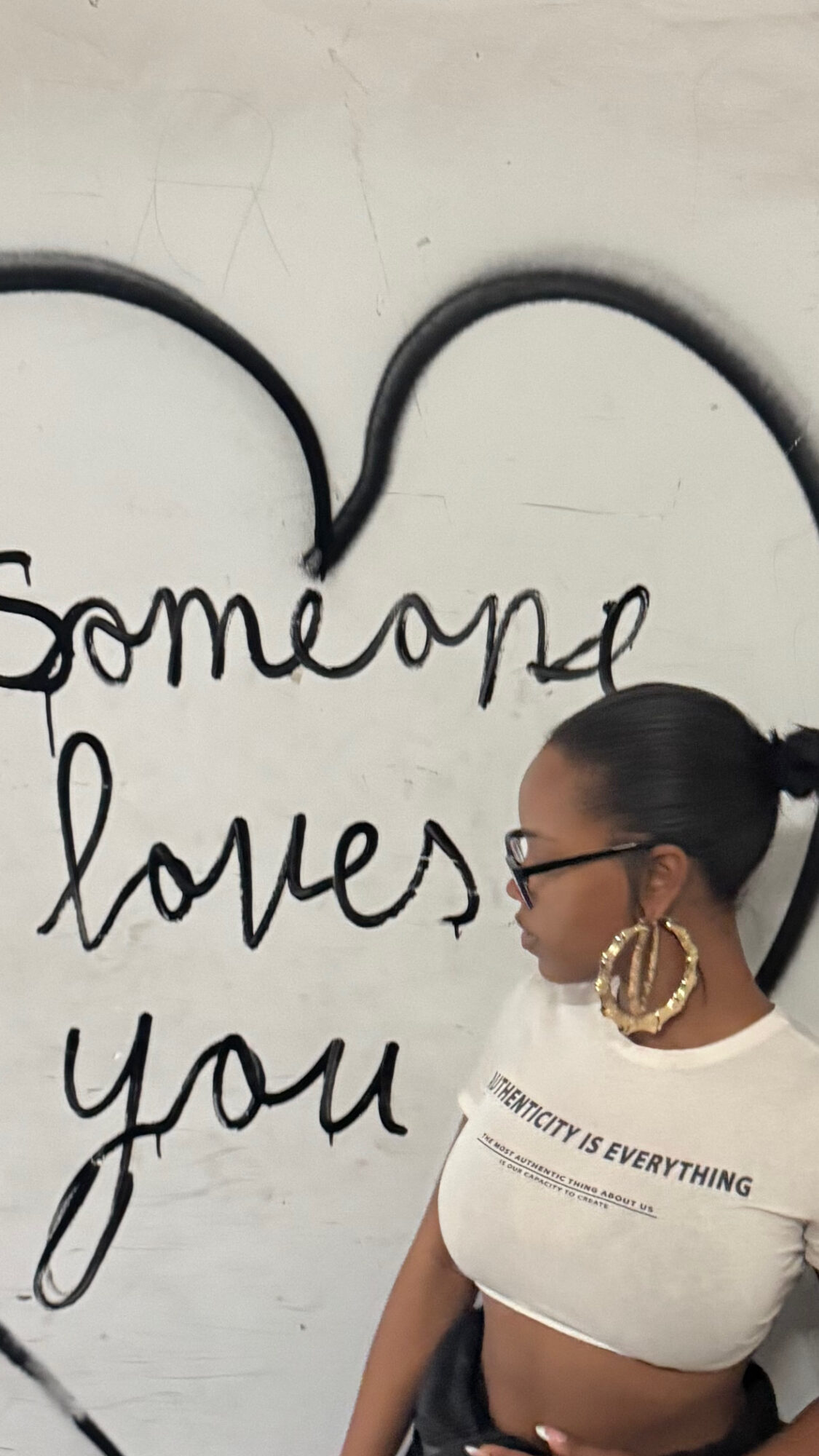 Woman with glasses and hoop earrings looking to the side, standing in front of a wall with handwritten text and a heart outline.