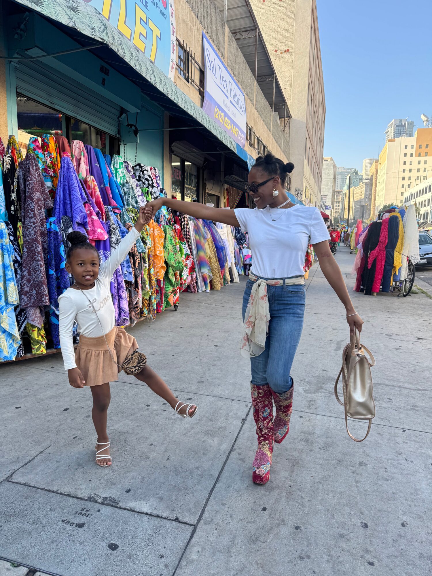 Two children standing on a city sidewalk near a clothing stall, one holding a bag, with buildings in the background.