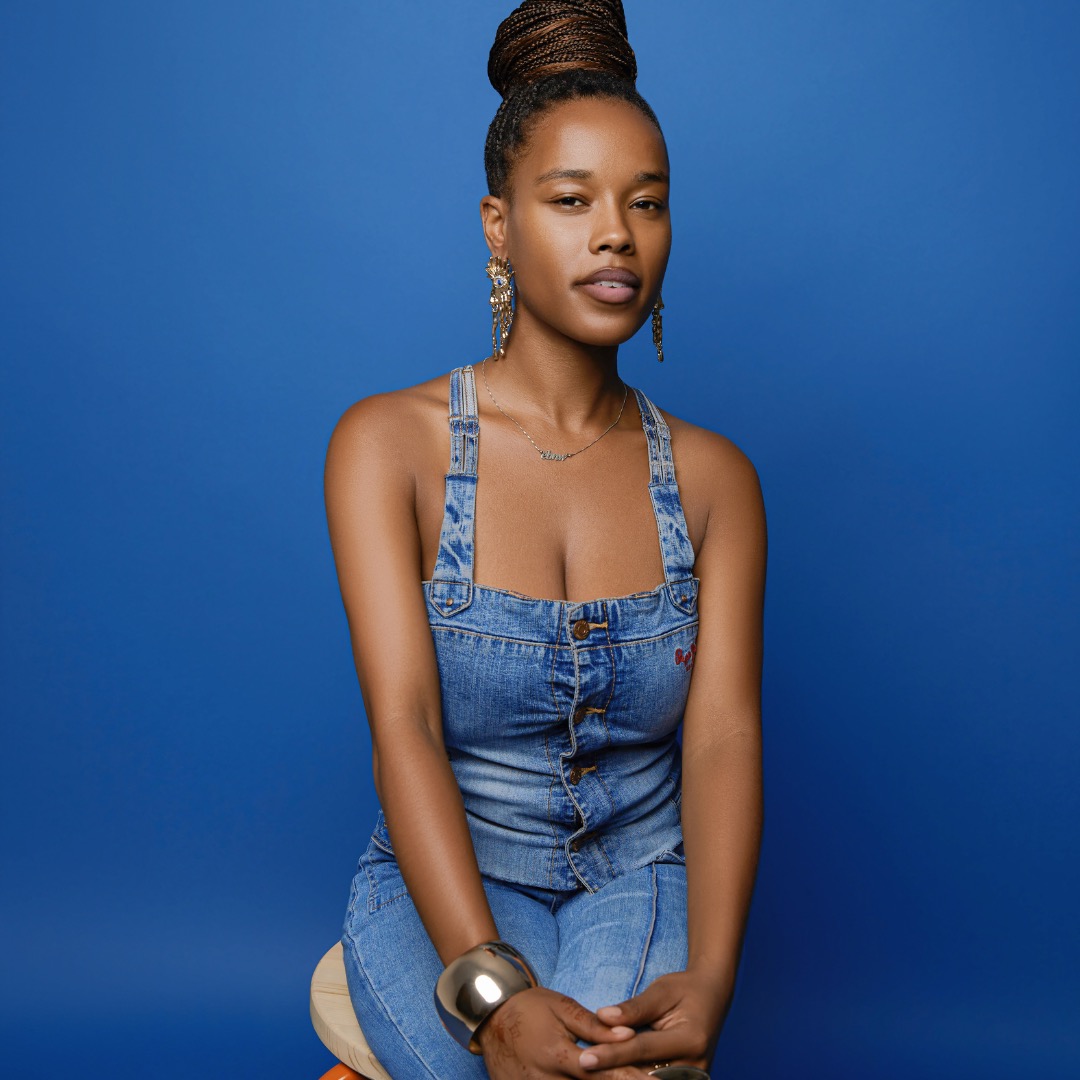 Young woman with braided hair in a denim outfit against a blue background, looking at the camera with a neutral expression.