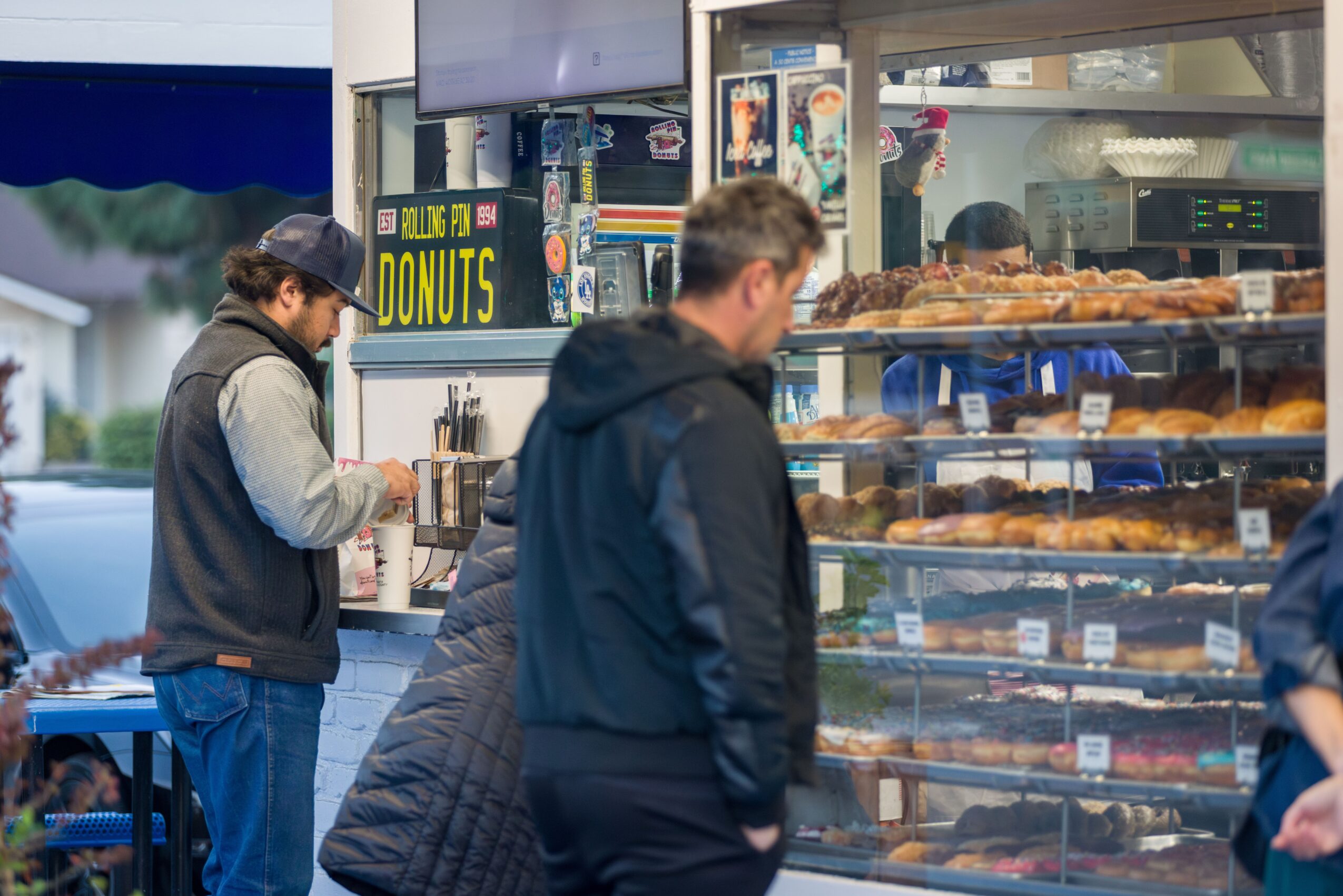 Two people stand outside a bakery with a display of baked goods, one person is looking at the display, another is using a phone.