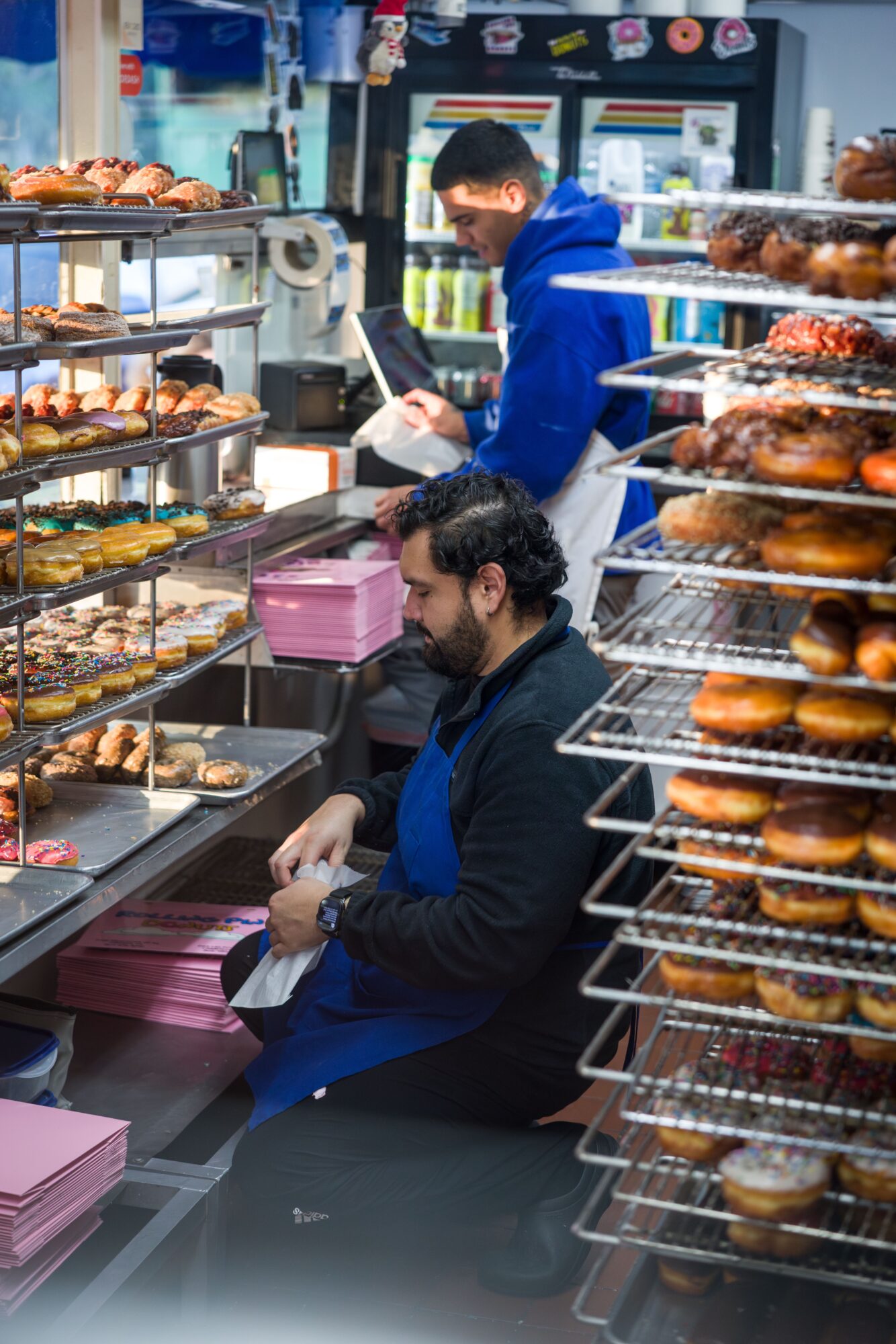 Two men working behind a bakery counter with shelves of baked goods, one sitting and one standing, in a bakery or cafe.