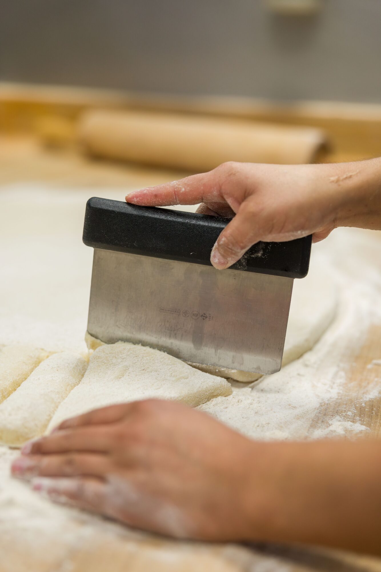 Person's hand presses dough with a bench scraper on a surface, with a rolling pin in the background.