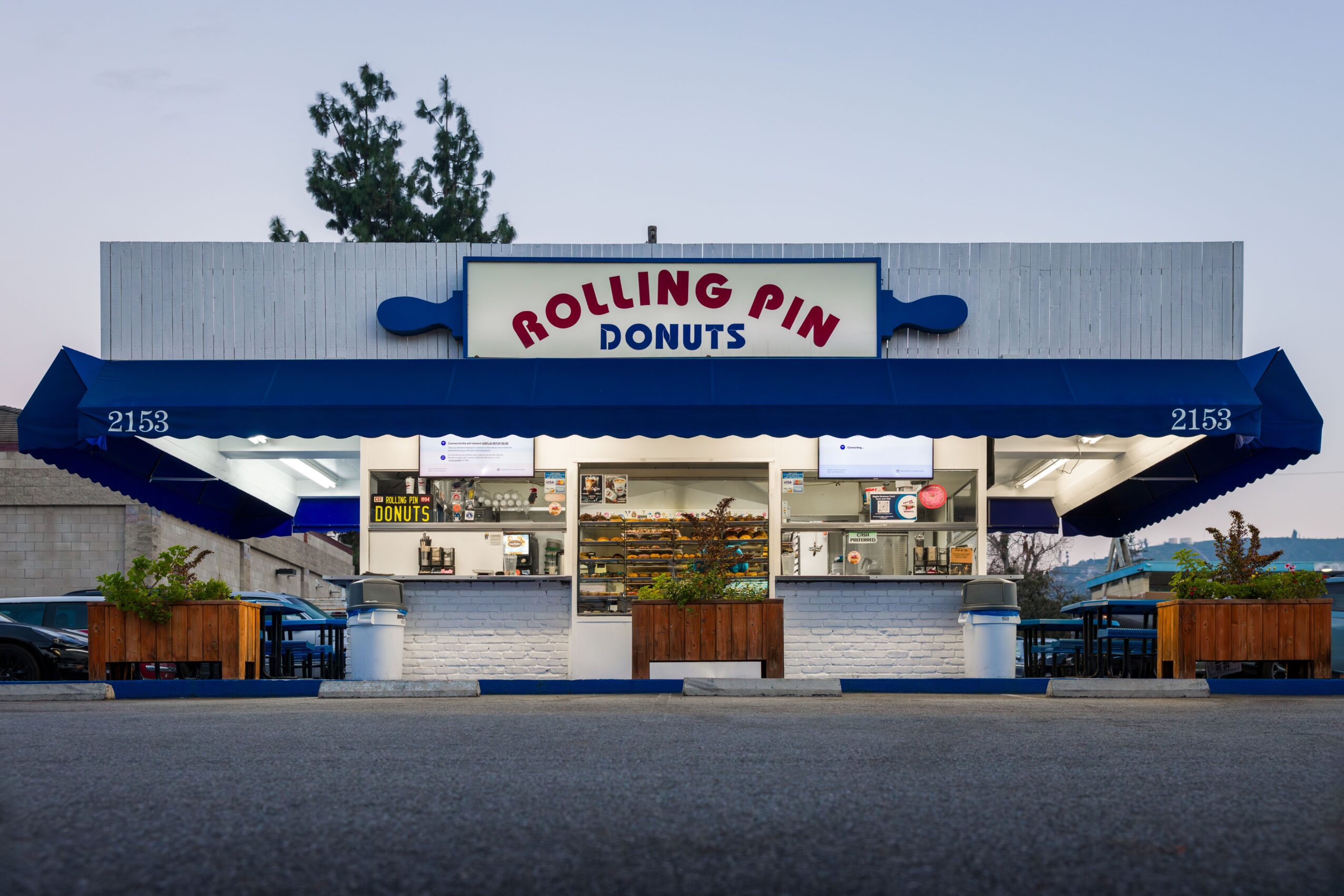 Donut shop with blue awning and sign reading 'Rolling Pin Donuts', outdoor seating, and planters, under a clear sky.