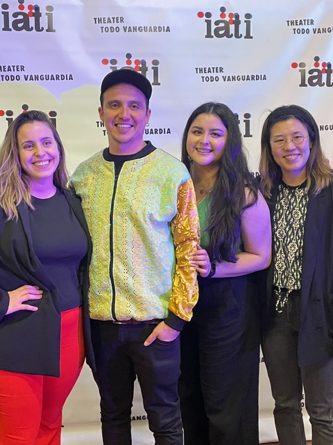 Four people standing in front of a backdrop with text, smiling, with one person wearing a yellow jacket and others in dark clothing.