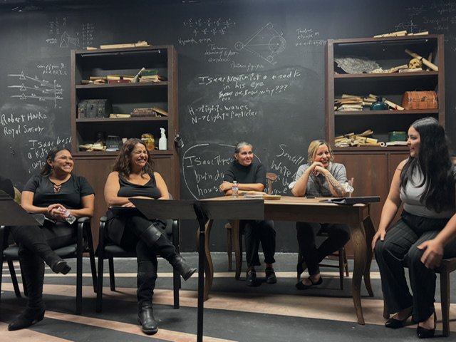 Five women sitting and standing around a table in a classroom with chalkboards and shelves filled with books and supplies.