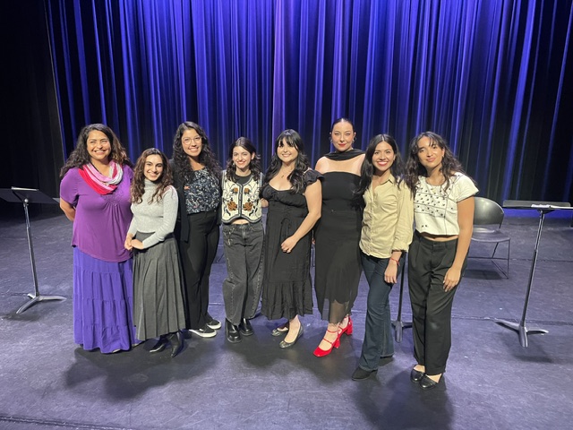 Group of eight women standing on stage with blue curtains behind them, smiling, dressed in various outfits.