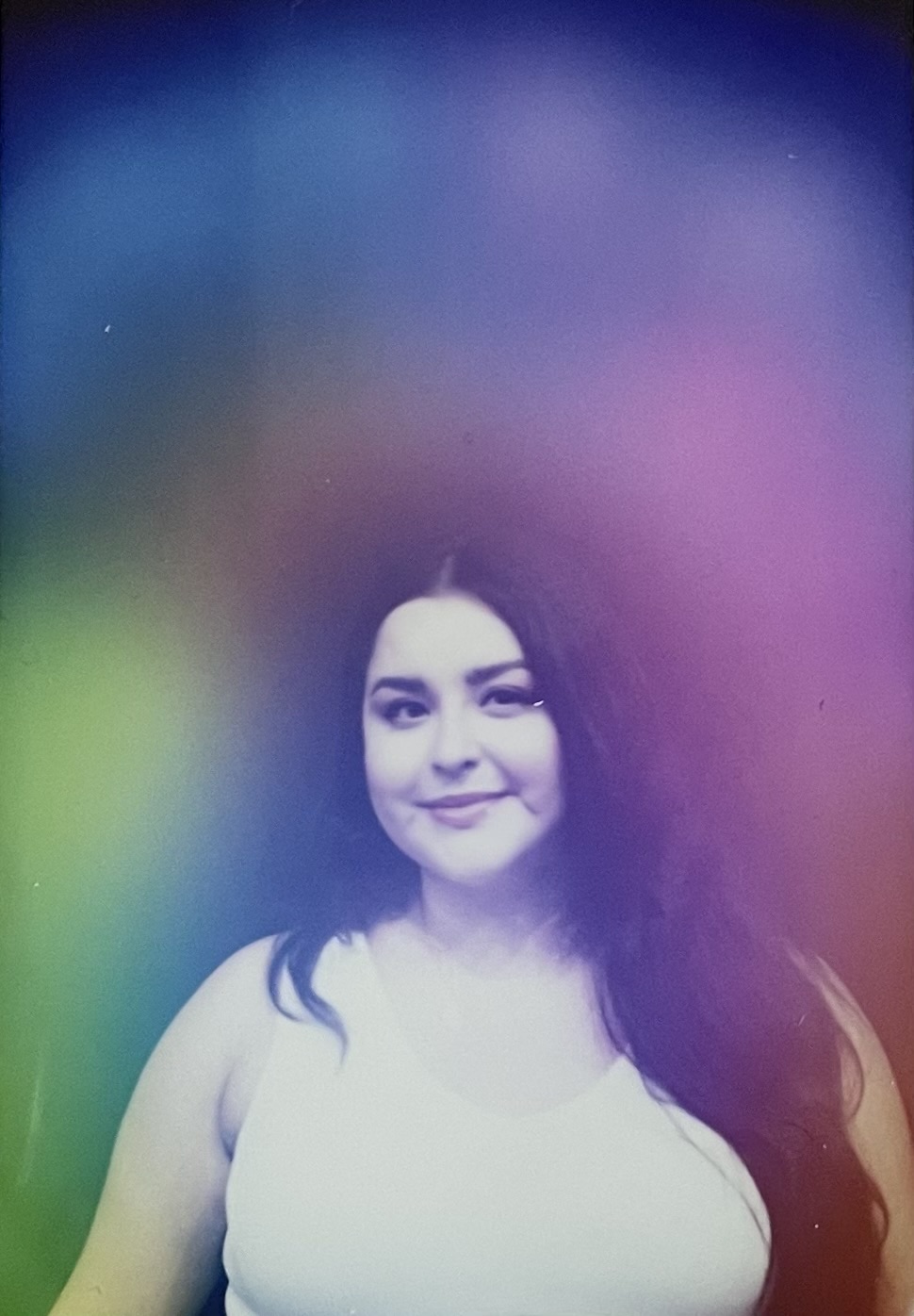 A woman with long dark hair smiling, wearing a sleeveless top, against a colorful blurred background.