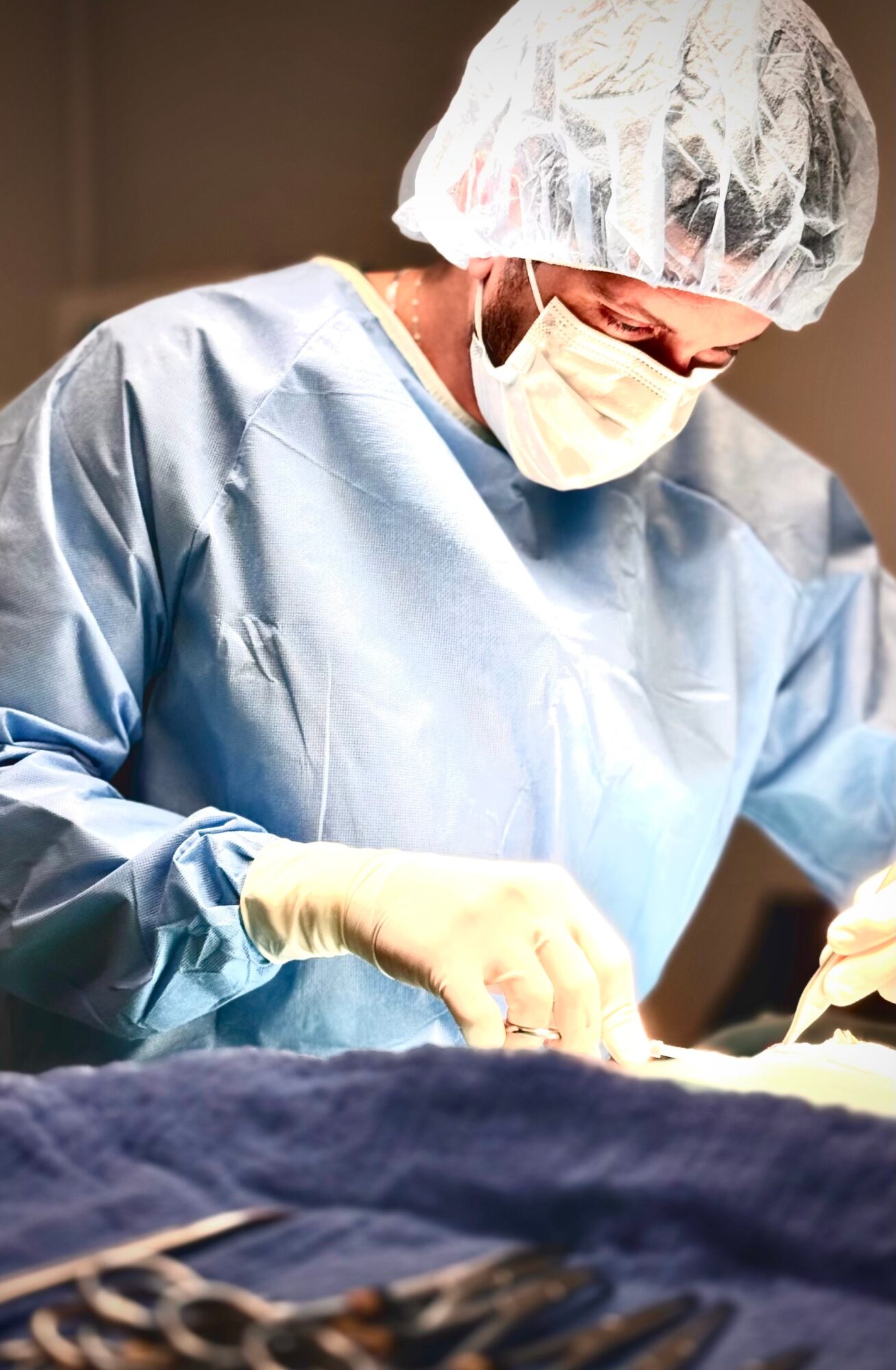 Medical professional wearing a surgical cap, mask, gown, and gloves performing a procedure in an operating room.