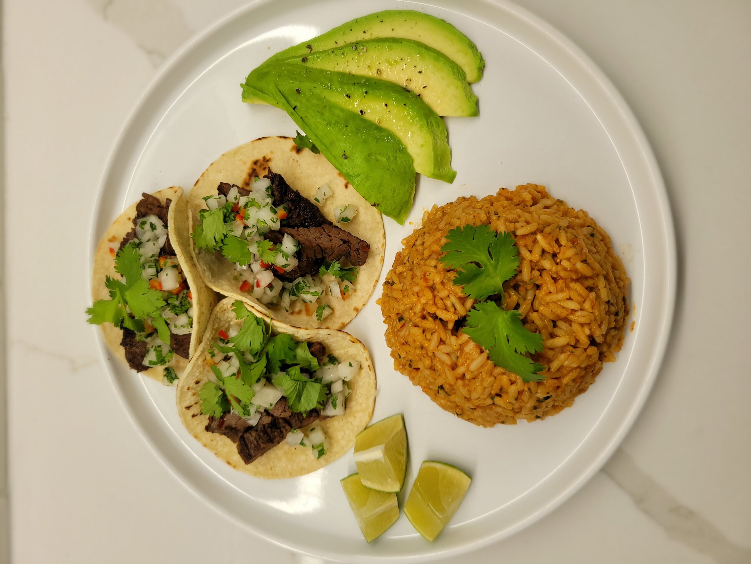 Three tacos with meat and cilantro, sliced avocado, a mound of rice with cilantro, and two lime wedges on a white plate.