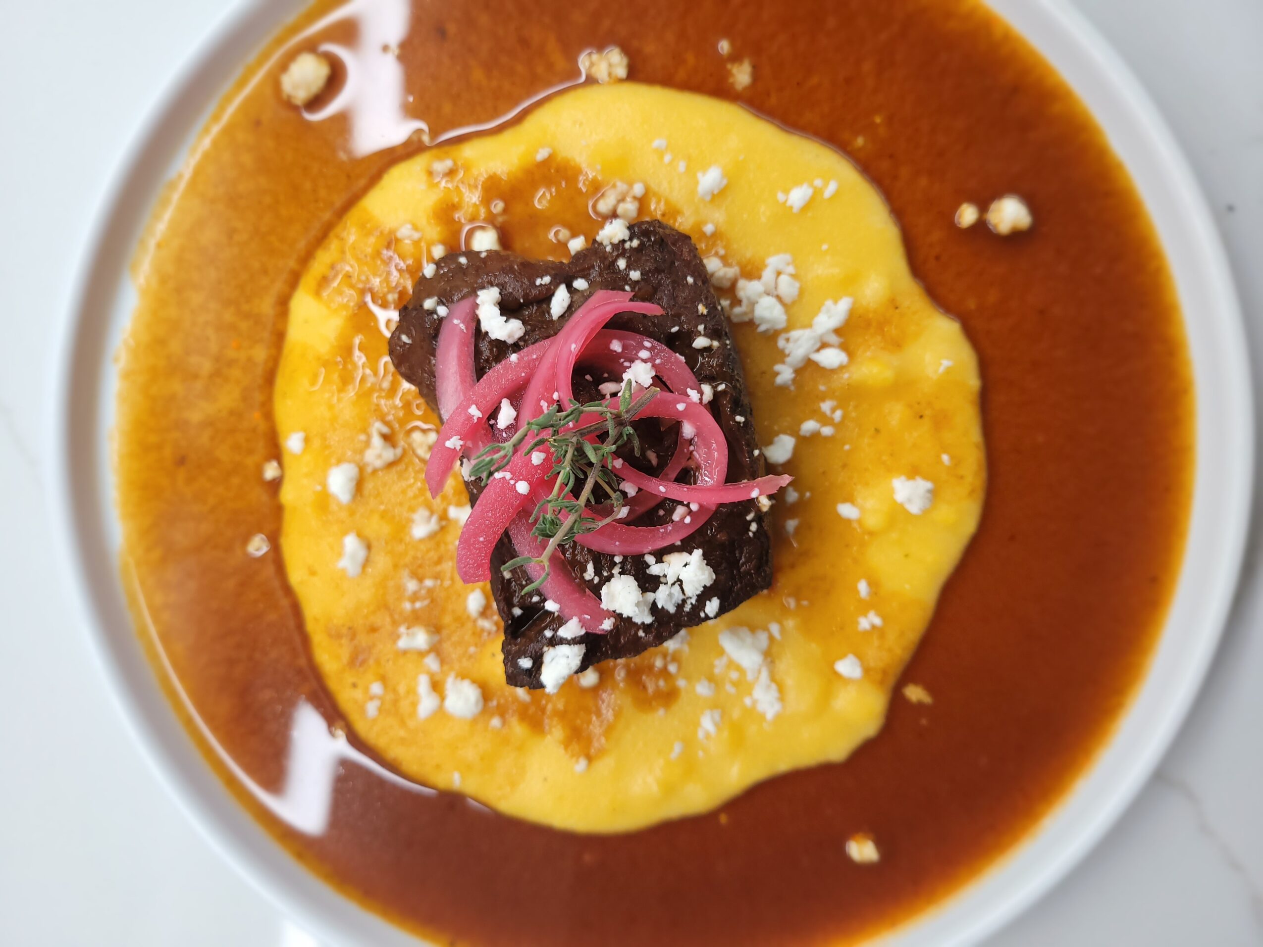A dish with a yellow base topped with dark sauce, pink pickled onions, and white crumbles, served in a white bowl.