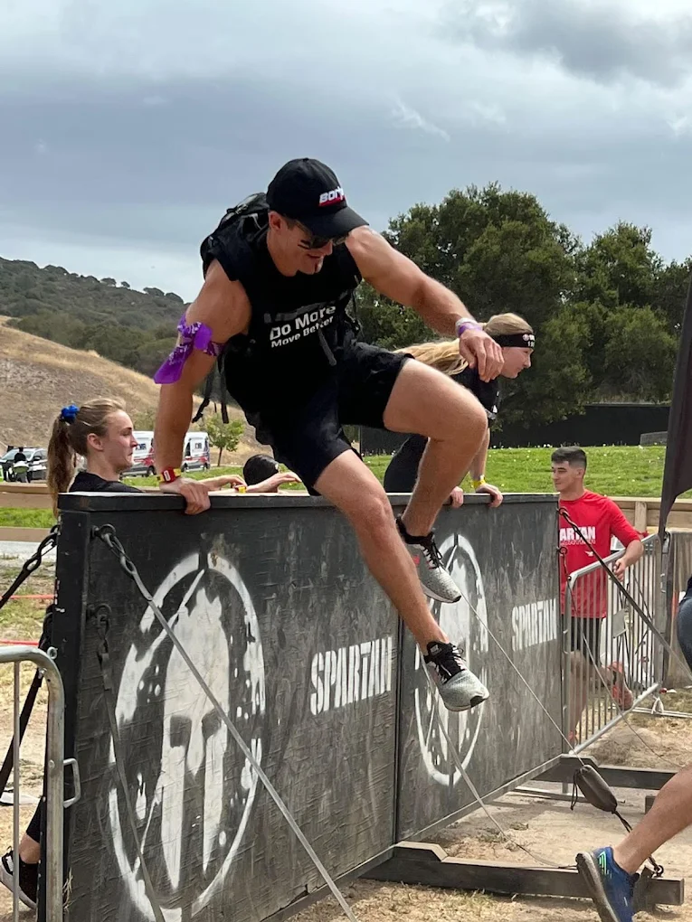 Person jumping over a barrier during an obstacle course, with spectators watching, outdoors on cloudy day.