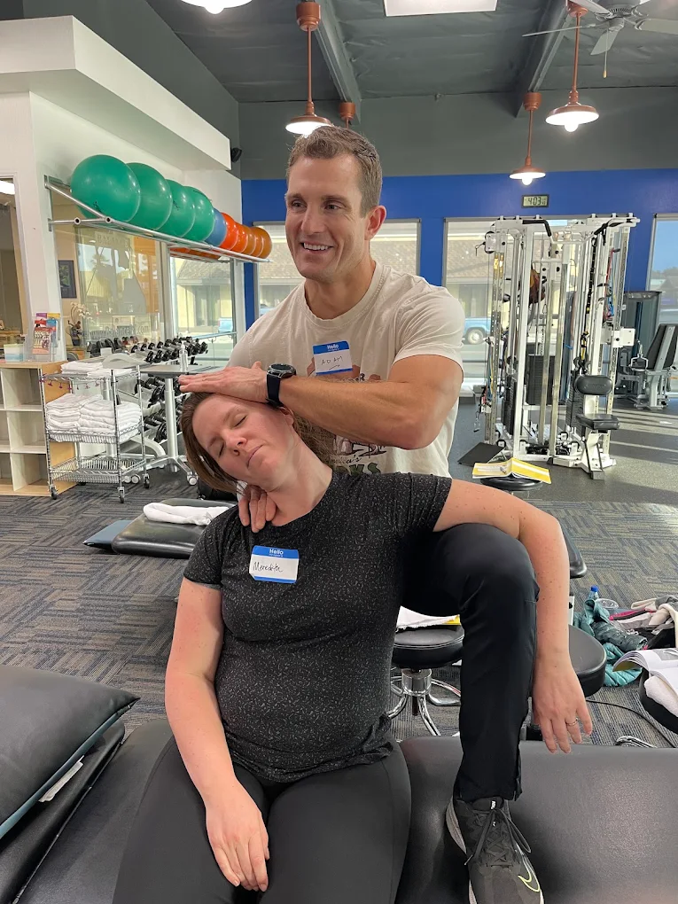 A man and woman in a gym, with the man standing behind the woman, smiling, and the woman sitting with her head tilted back.