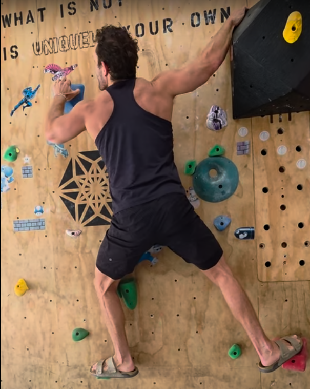 Person climbing an indoor bouldering wall with colorful holds, facing away, wearing a black tank top and shorts.