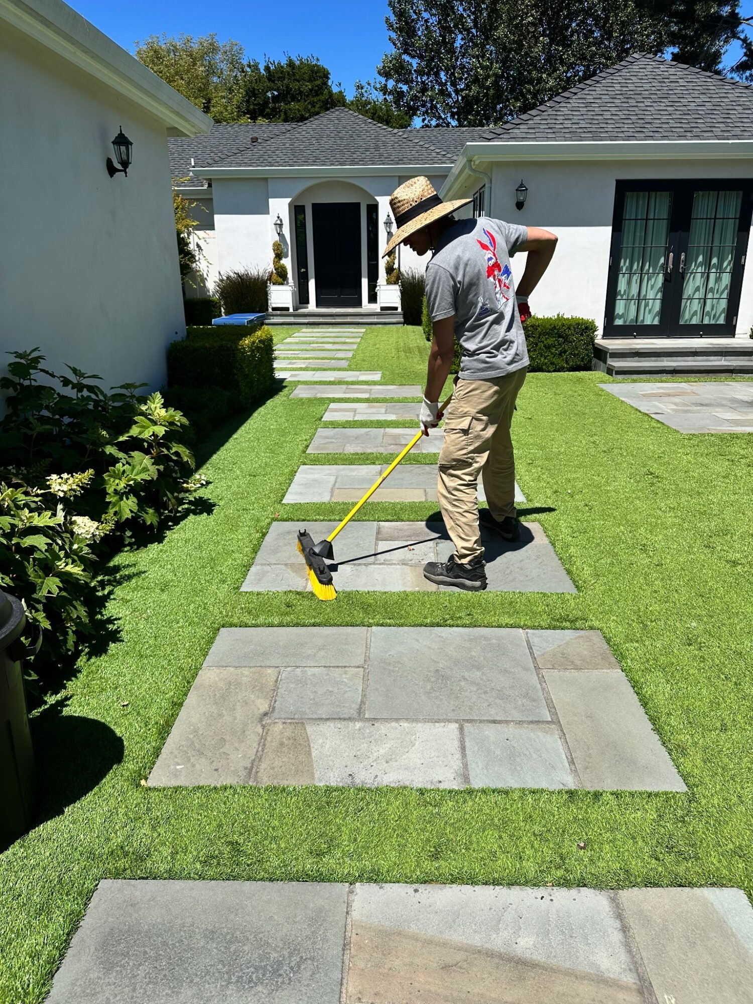 Person installing or maintaining a stone pathway in a backyard with green grass and white houses, wearing a hat and casual clothes.