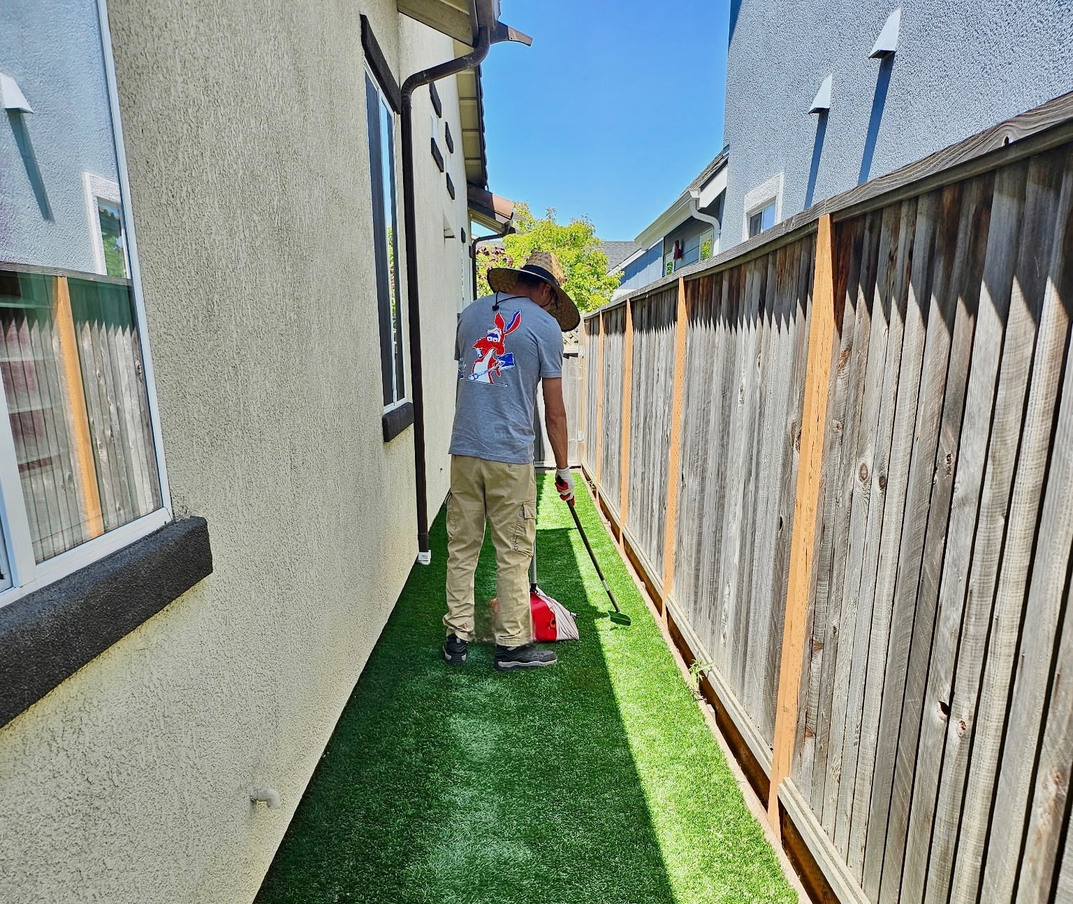 Child wearing a hat and blue shirt mows a narrow backyard with artificial grass, fence on the right, house on the left, sunny sky.