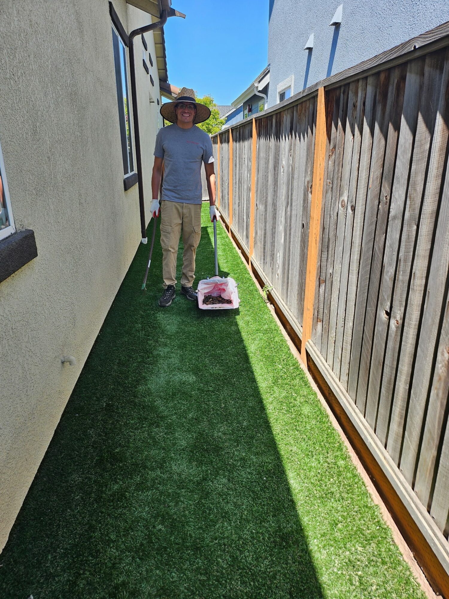 Person wearing a hat and gloves pushes a lawn mower along a narrow grassy path between two fences.