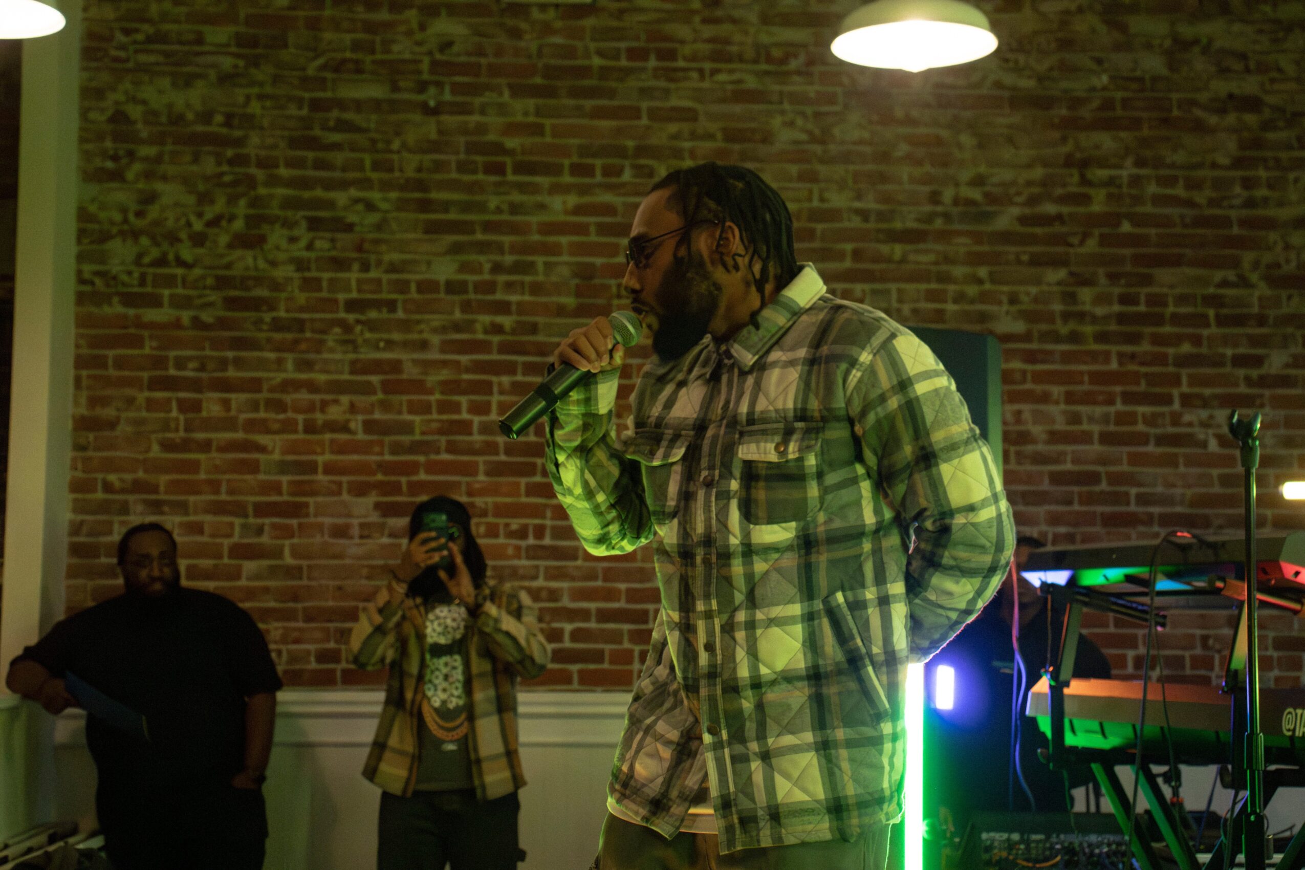 Person with braided hair and glasses singing into a microphone on stage, brick wall background, stage lighting