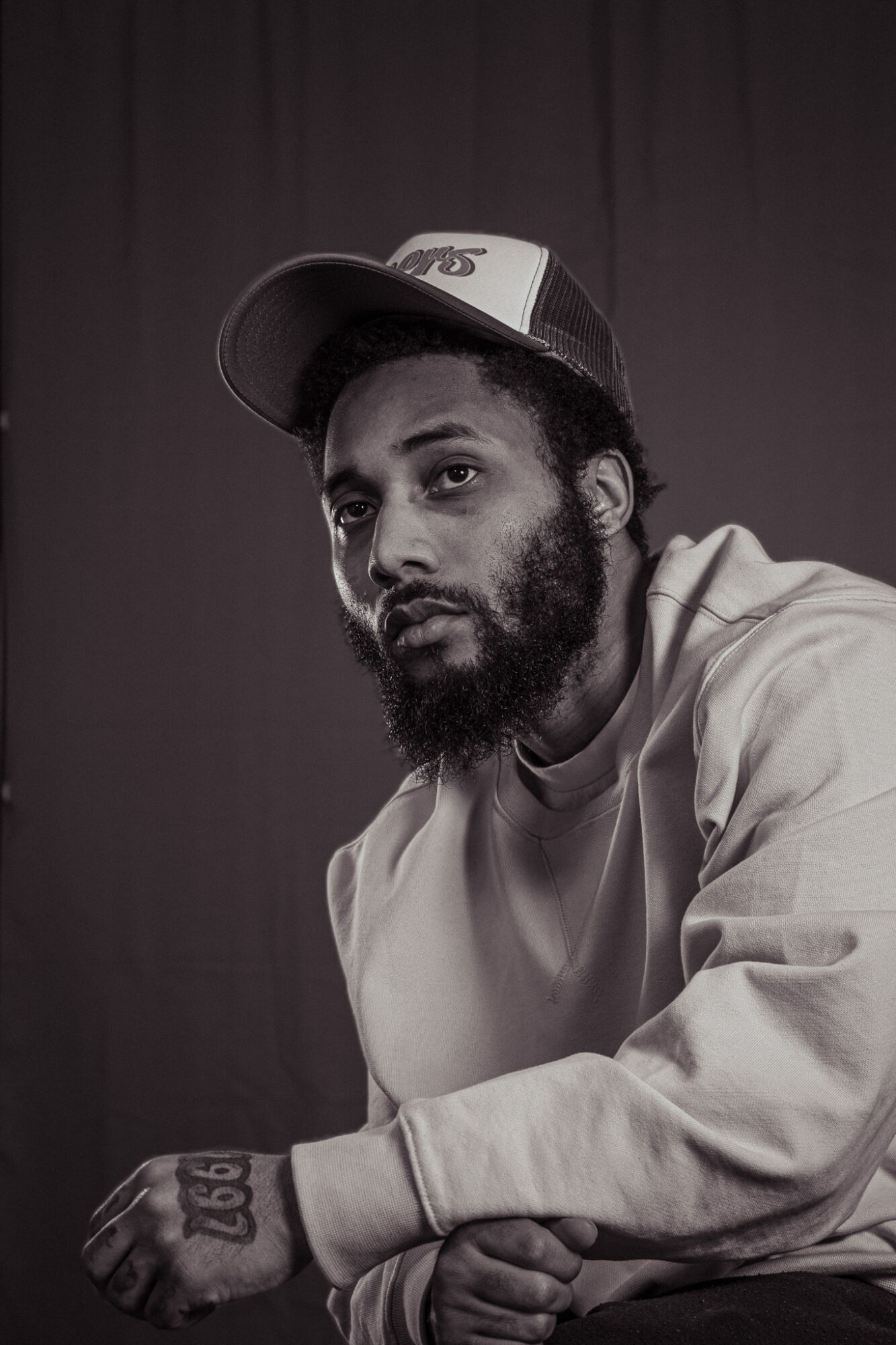 Man with beard wearing a cap and sweatshirt, sitting against a dark background.