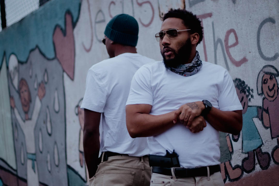 Two men standing outdoors near a colorful mural, one facing away and the other looking to the side, wearing casual clothing.