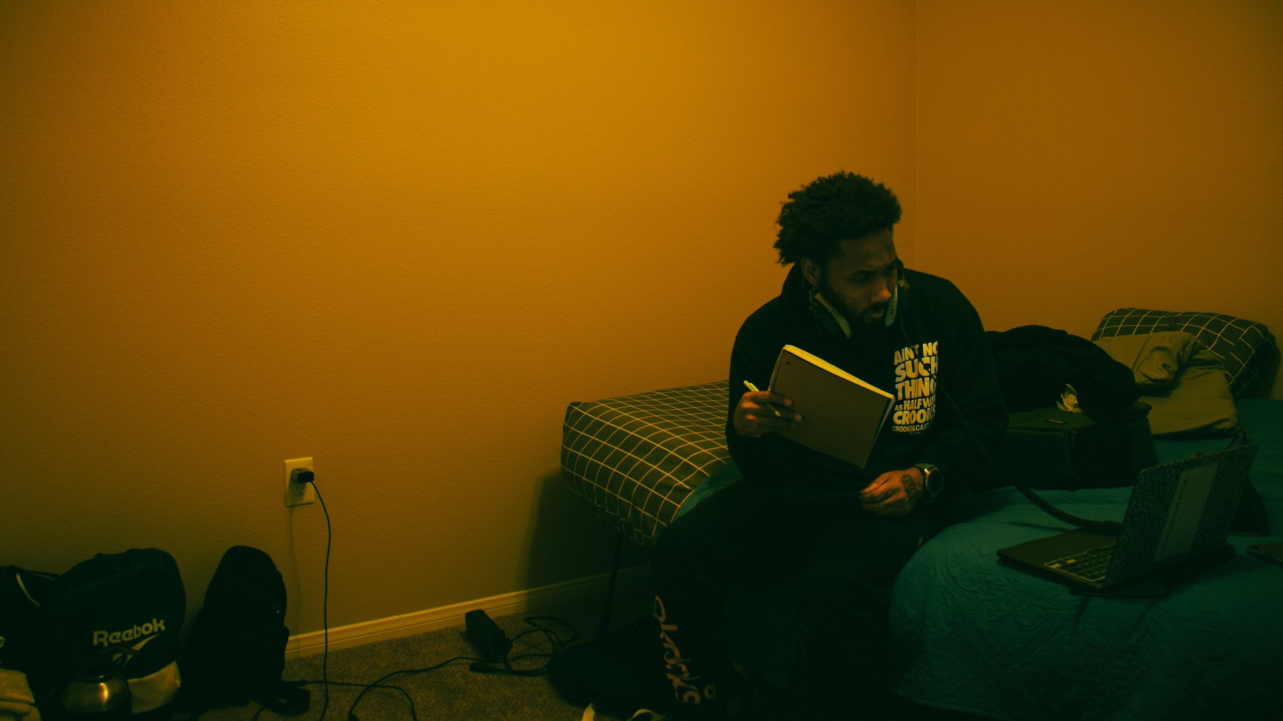 Person sitting on bed reading a book in a dimly lit room with yellow walls.