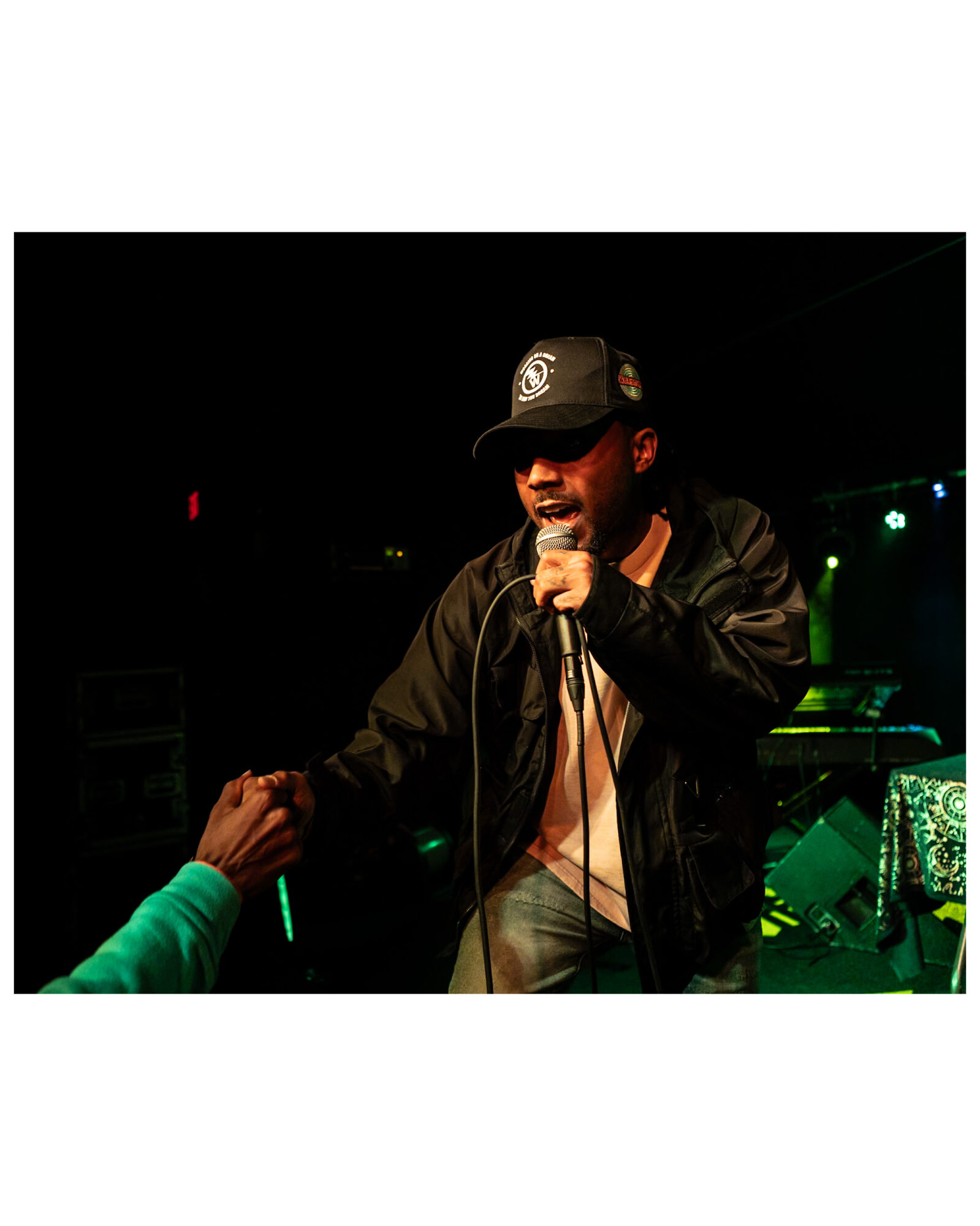 Man performing on stage, holding microphone, wearing a black jacket and cap, engaging with audience in a dark setting.