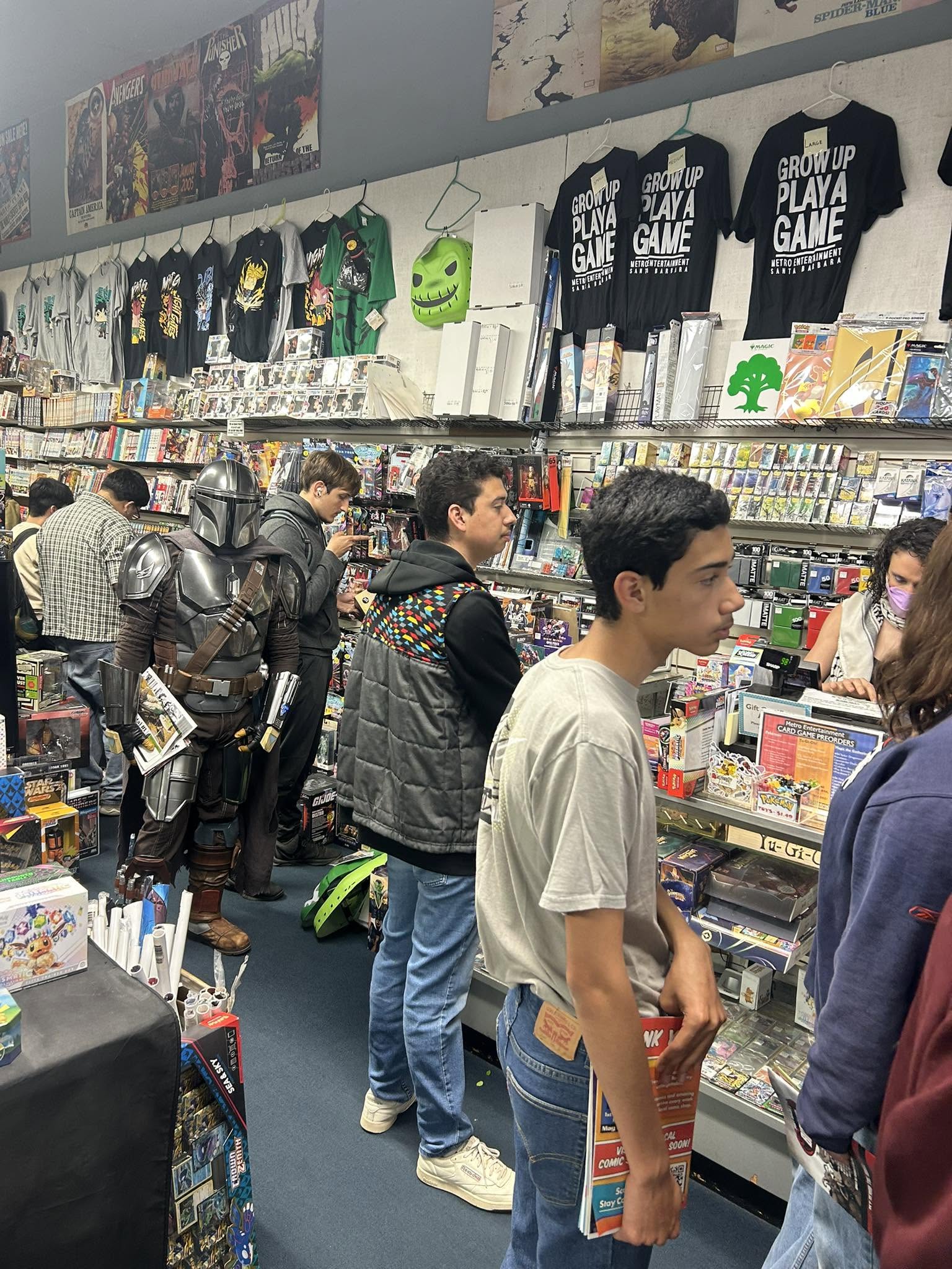 People shopping in a store aisle with shelves filled with toys, games, and merchandise, some browsing and others waiting in line.