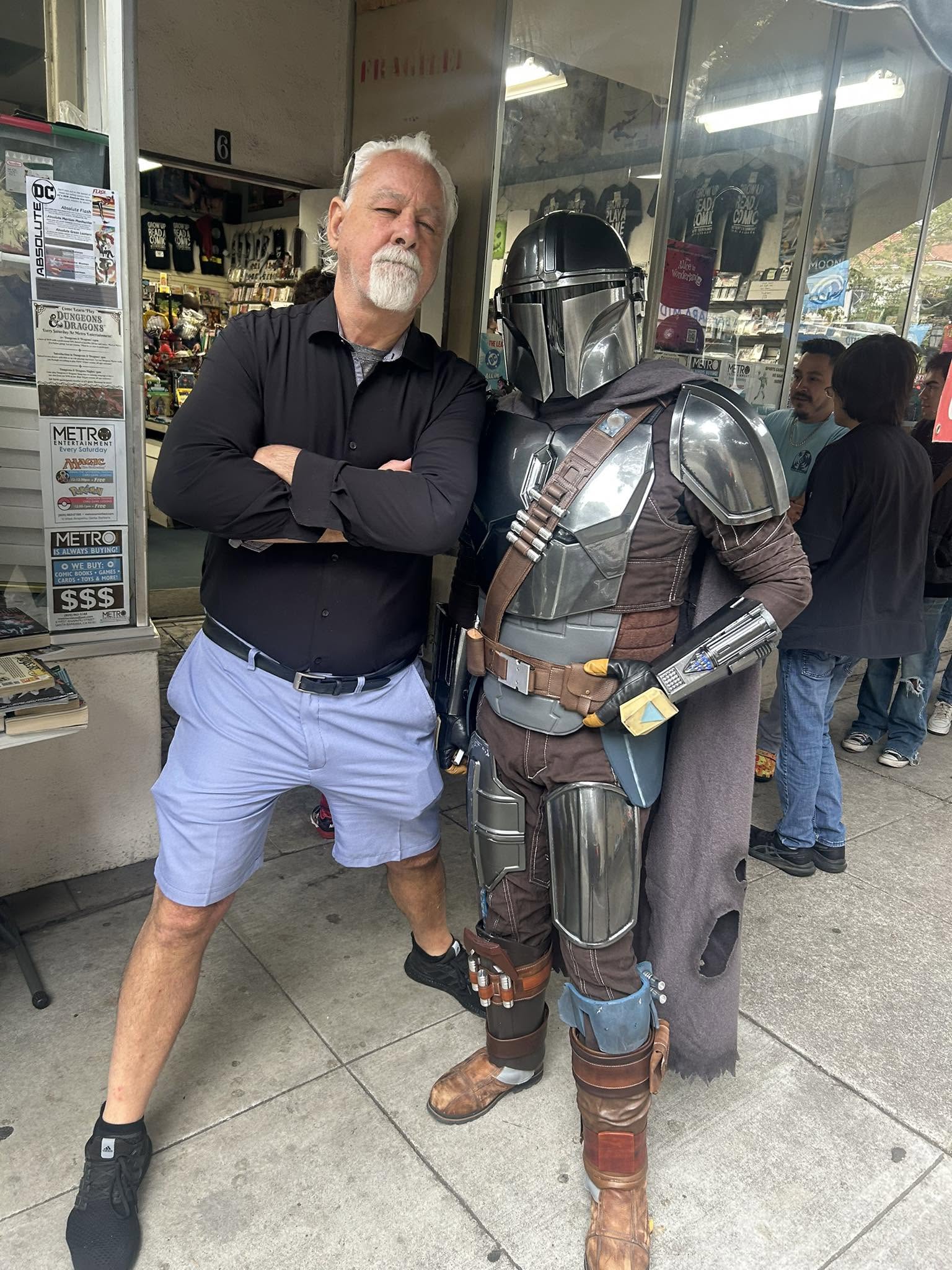 Two people standing outside a store, one in casual clothing and the other in Mandalorian costume, posing together.
