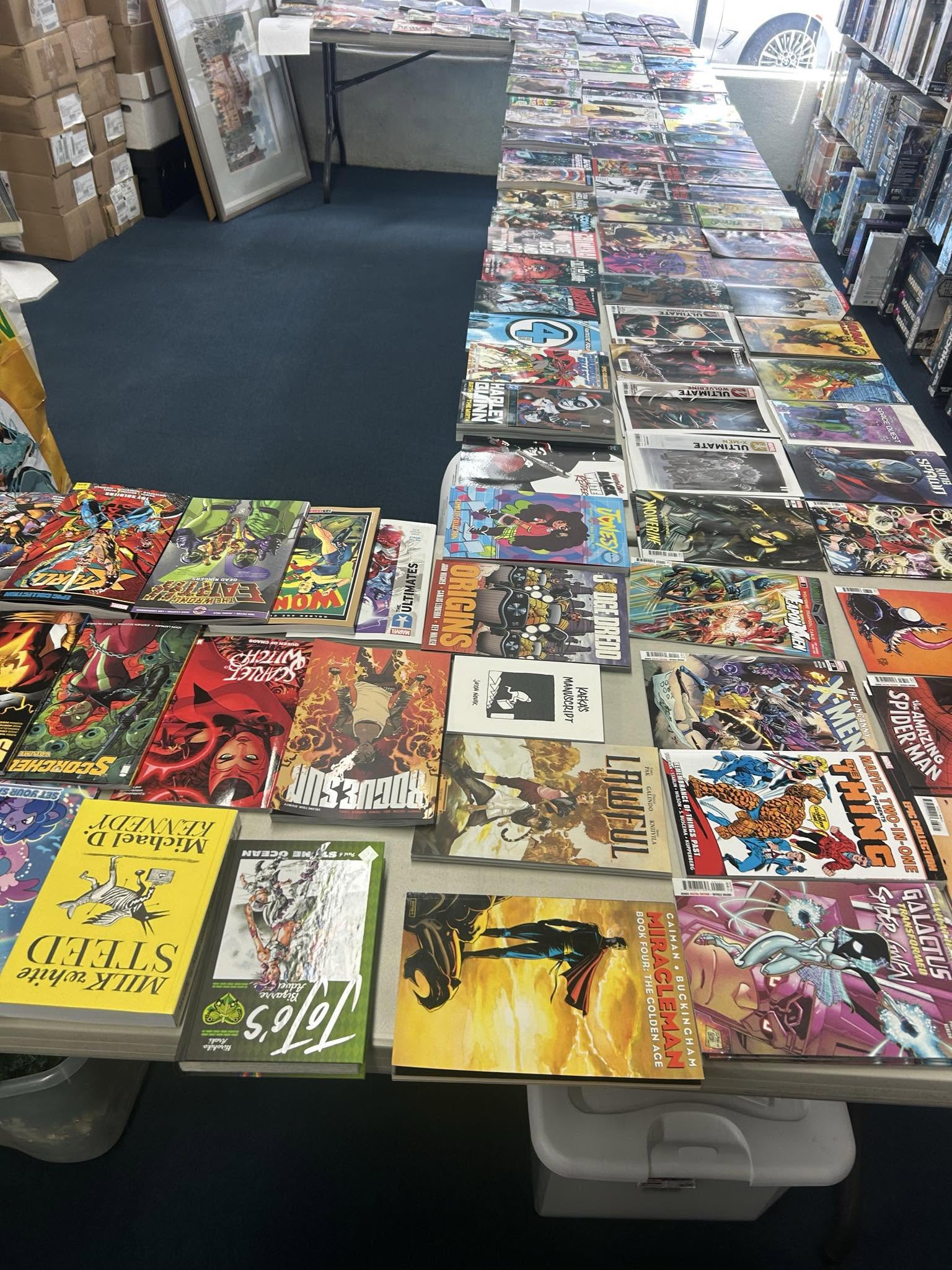 Table covered with comic books and graphic novels in a store, with shelves and boxes in the background.
