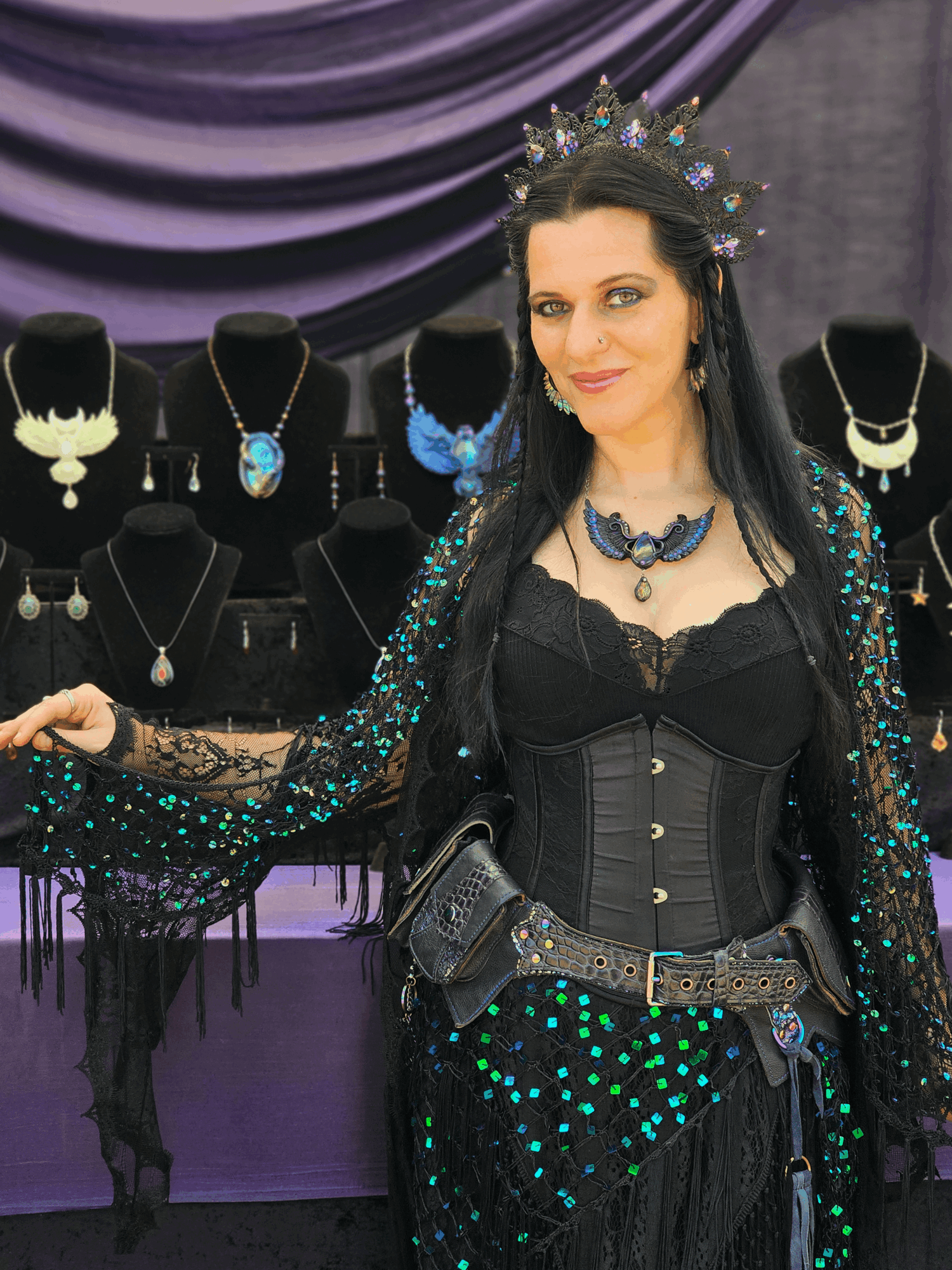 Woman dressed as a queen or princess with jewelry and a crown, standing in front of jewelry displays, smiling.