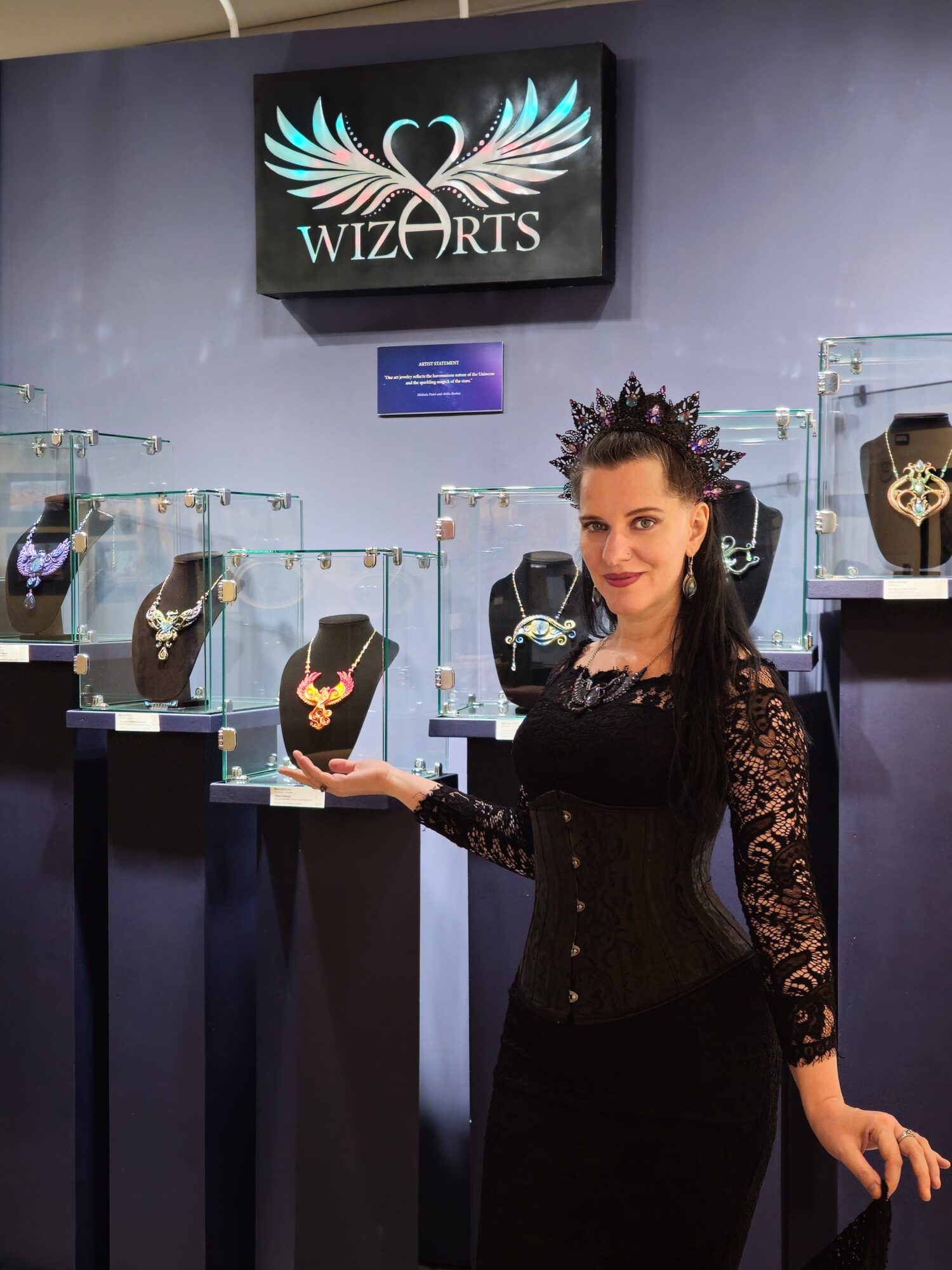 Woman with dark hair and crown displays jewelry in a store with a WIZARTS sign above.