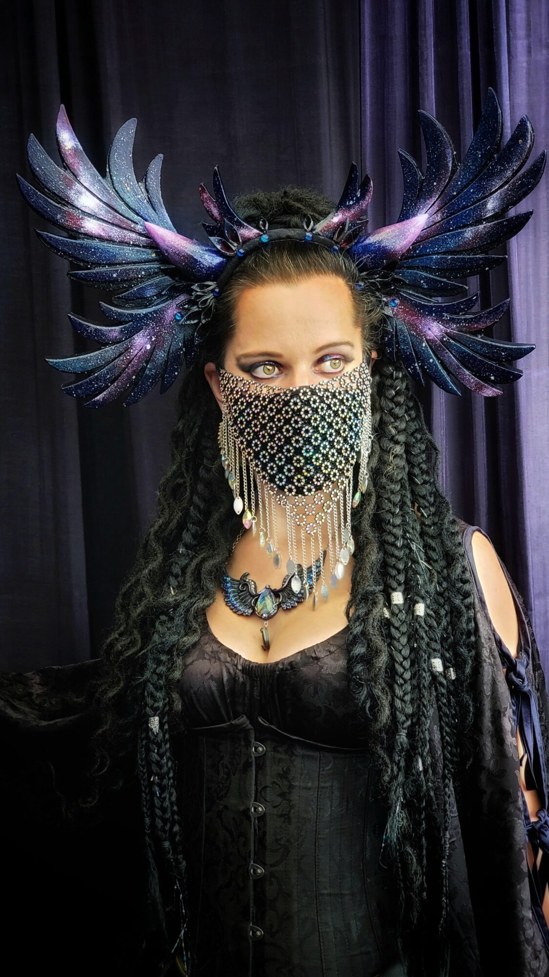 Person wearing an elaborate headpiece with feathers and a beaded mask, dressed in dark clothing, against a dark background.