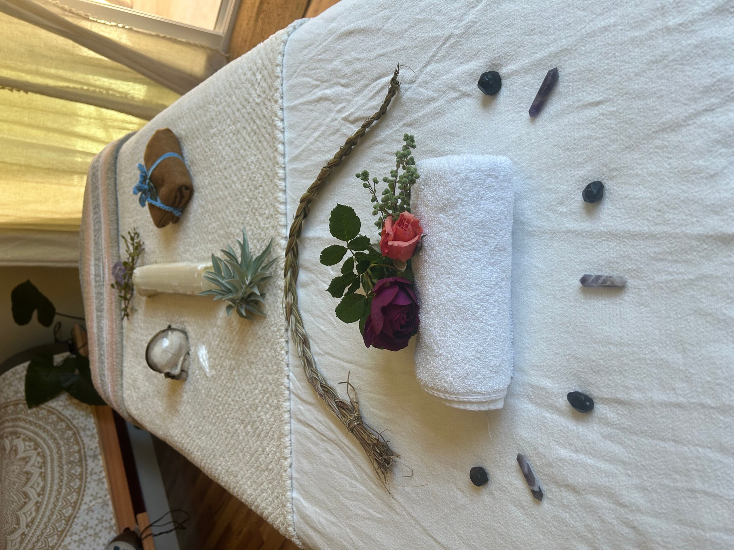 Spa setup with rolled towel, flowers, stones, and candles on a white towel, viewed from the side.