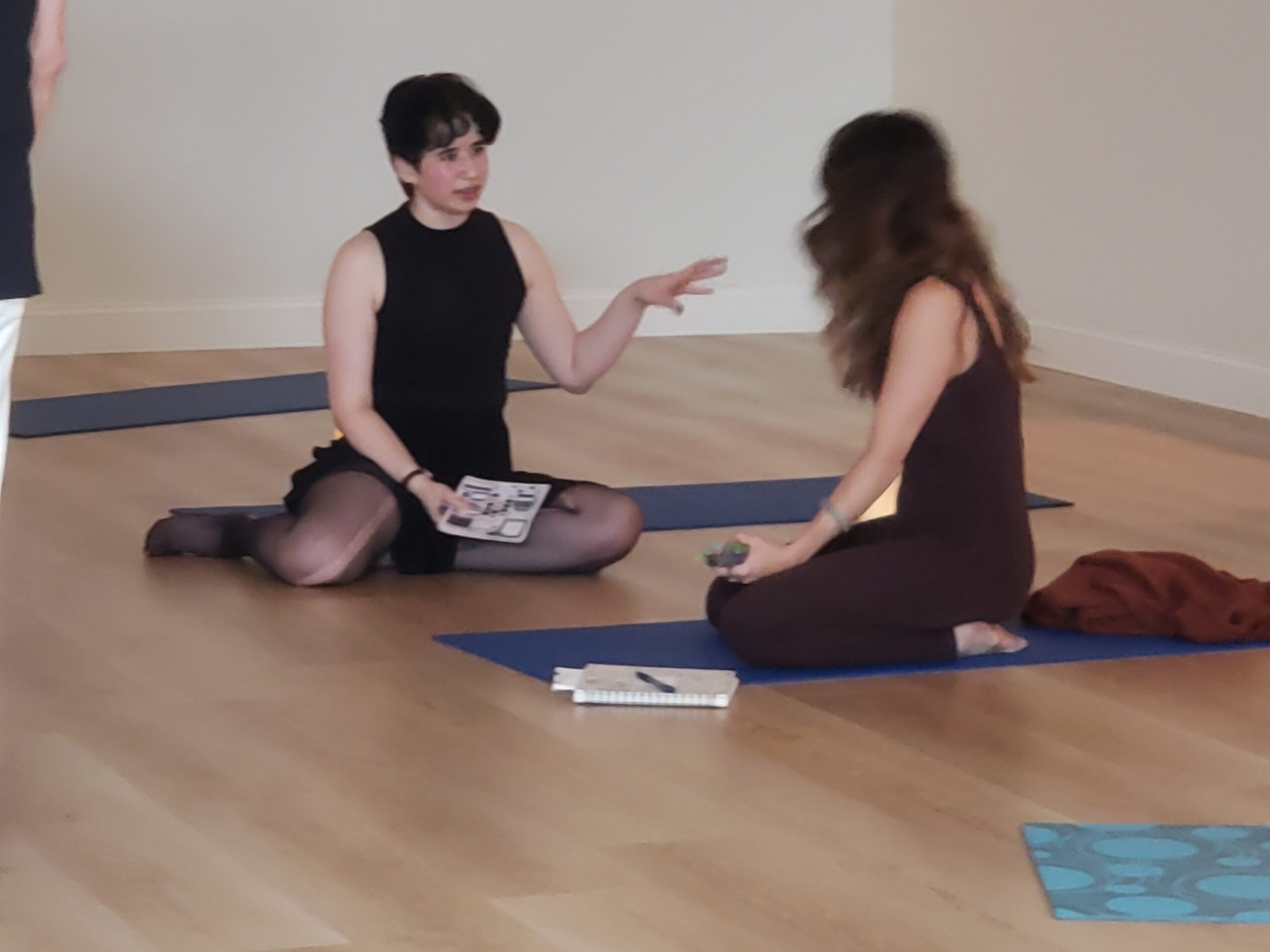 Two women sitting on yoga mats on a wooden floor, one gesturing with her hand, the other listening, with a notebook nearby.