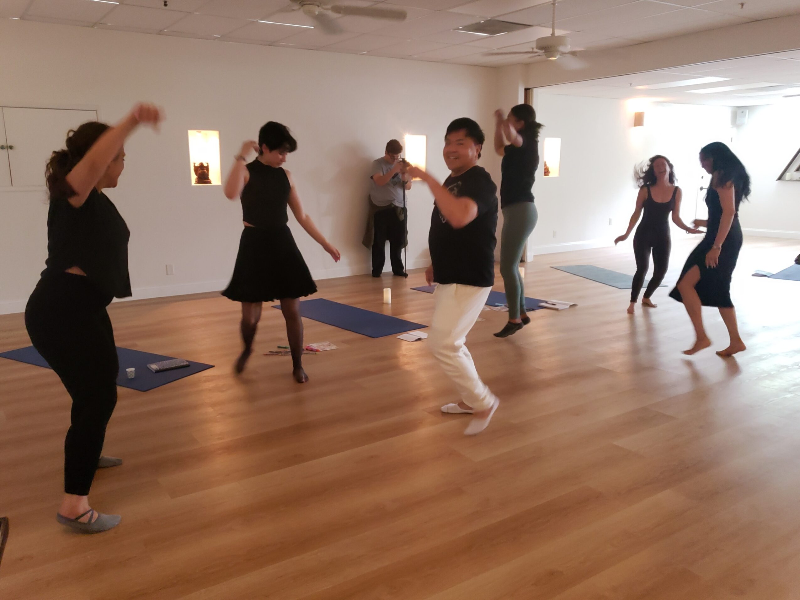 People dancing in a room with wooden floor and white walls, some with arms raised, others moving around.