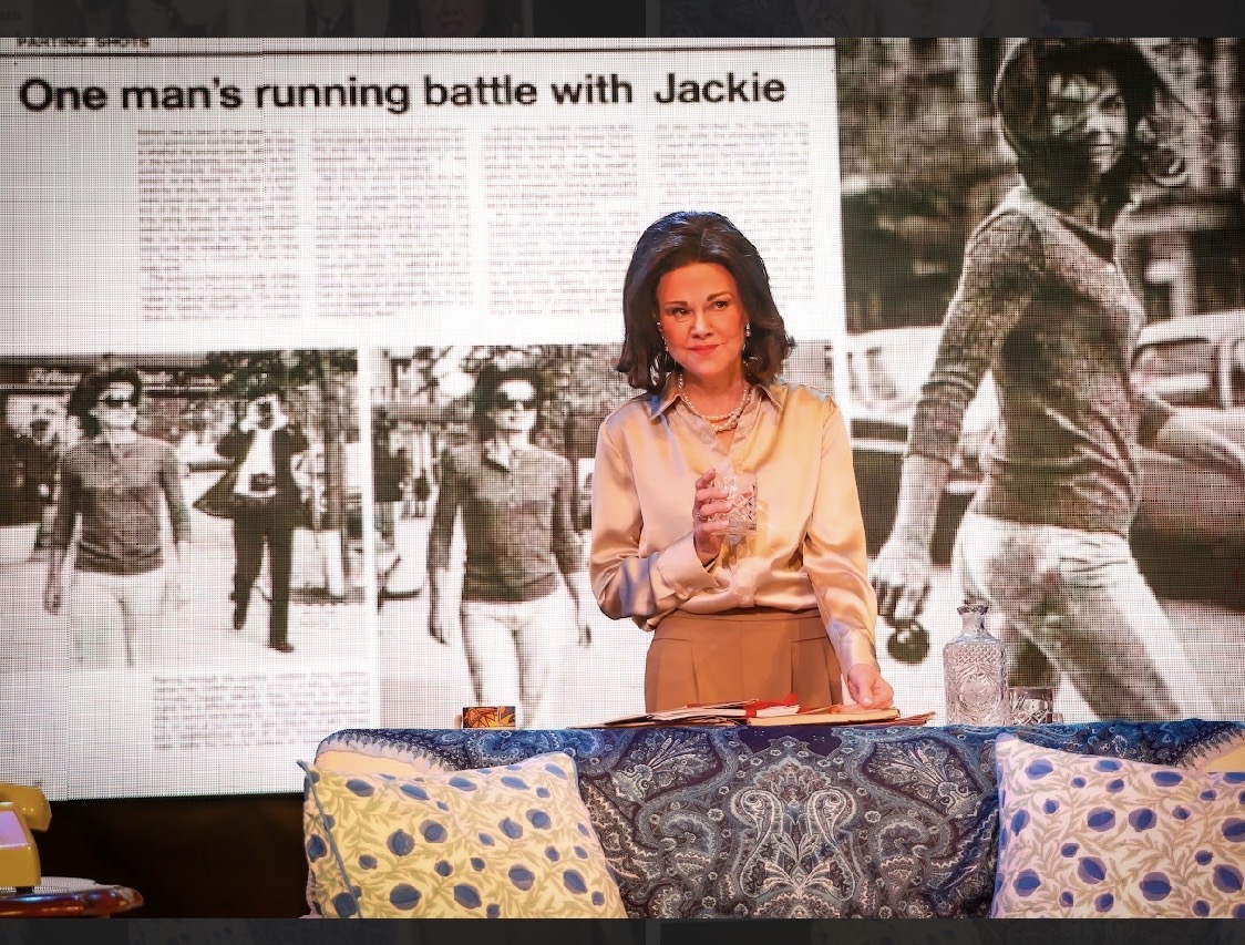 Woman standing in front of a newspaper article and black-and-white photos, with colorful cushions in foreground.