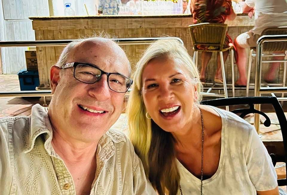 Two smiling people sitting together indoors, with a wooden counter and other people in the background.