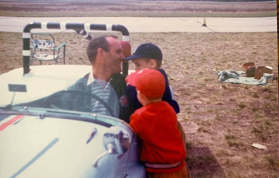 A man and two children near a small vehicle outdoors, with a goalpost and scattered objects in the background.