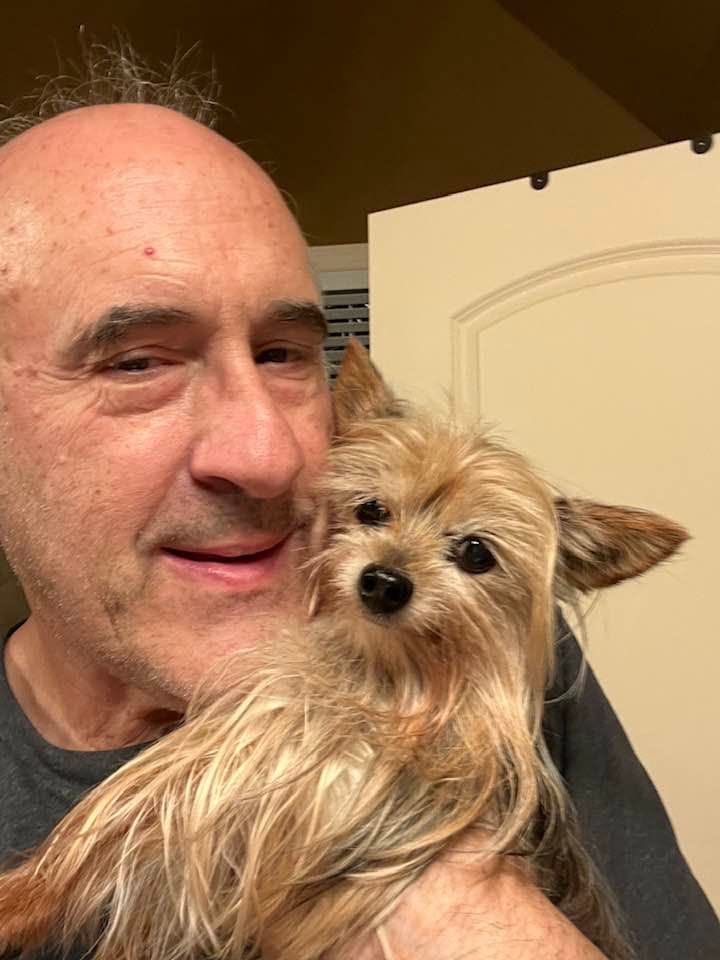 A man holding a small, fluffy dog close to his face indoors, with a neutral background.