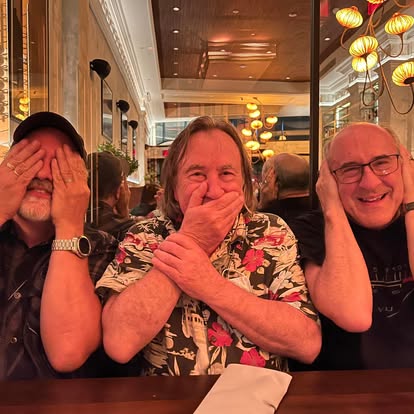 Three men sitting at a table, laughing and covering their faces, in a warmly lit restaurant or cafe.