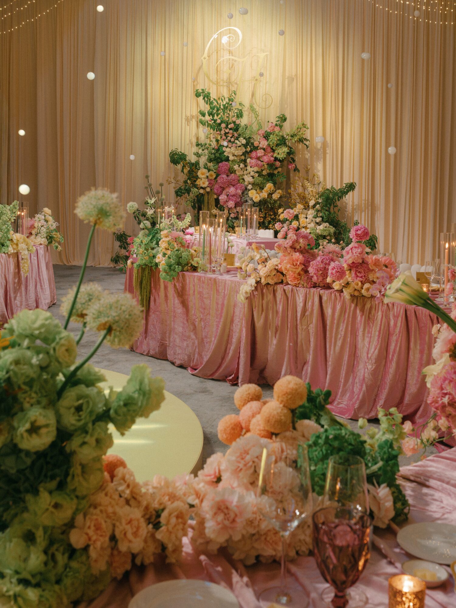 Decorated table with pink flowers and candles, backdrop with curtains and hanging lights, soft lighting, elegant event setting.