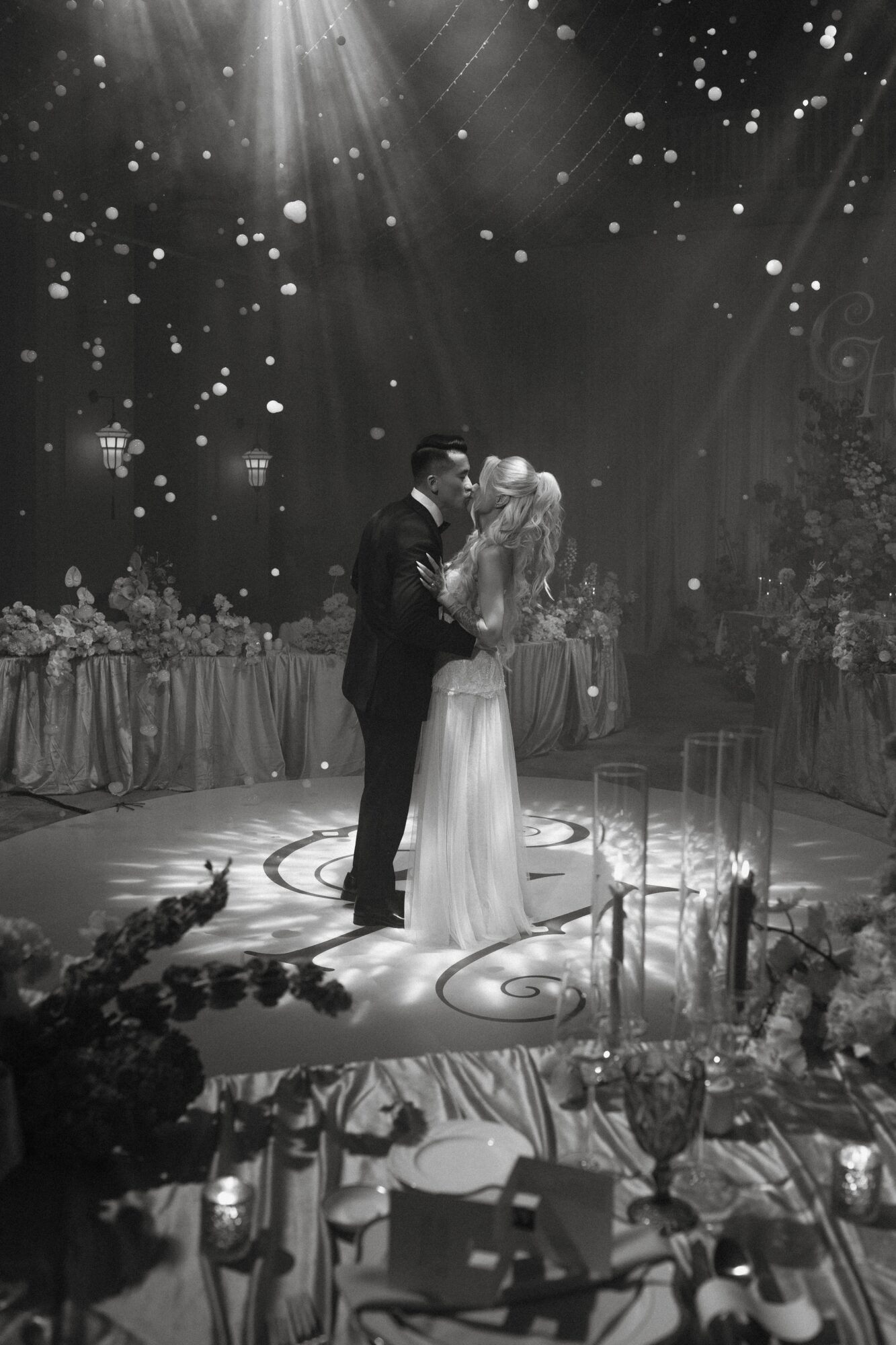 Couple dancing on a decorated stage with hanging lights and floral arrangements, surrounded by tables and candles.