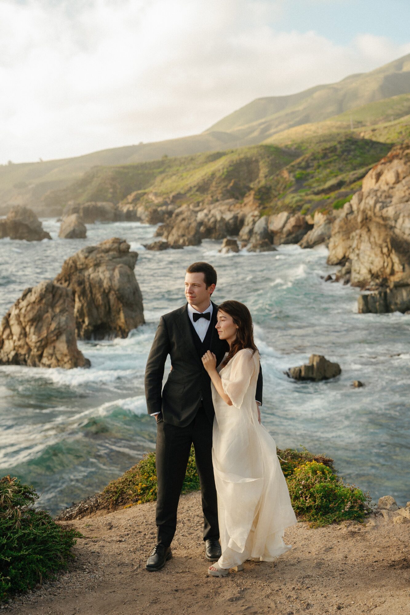 A couple in wedding attire stands on a rocky coastline with waves crashing behind them, overlooking hills and a cloudy sky.